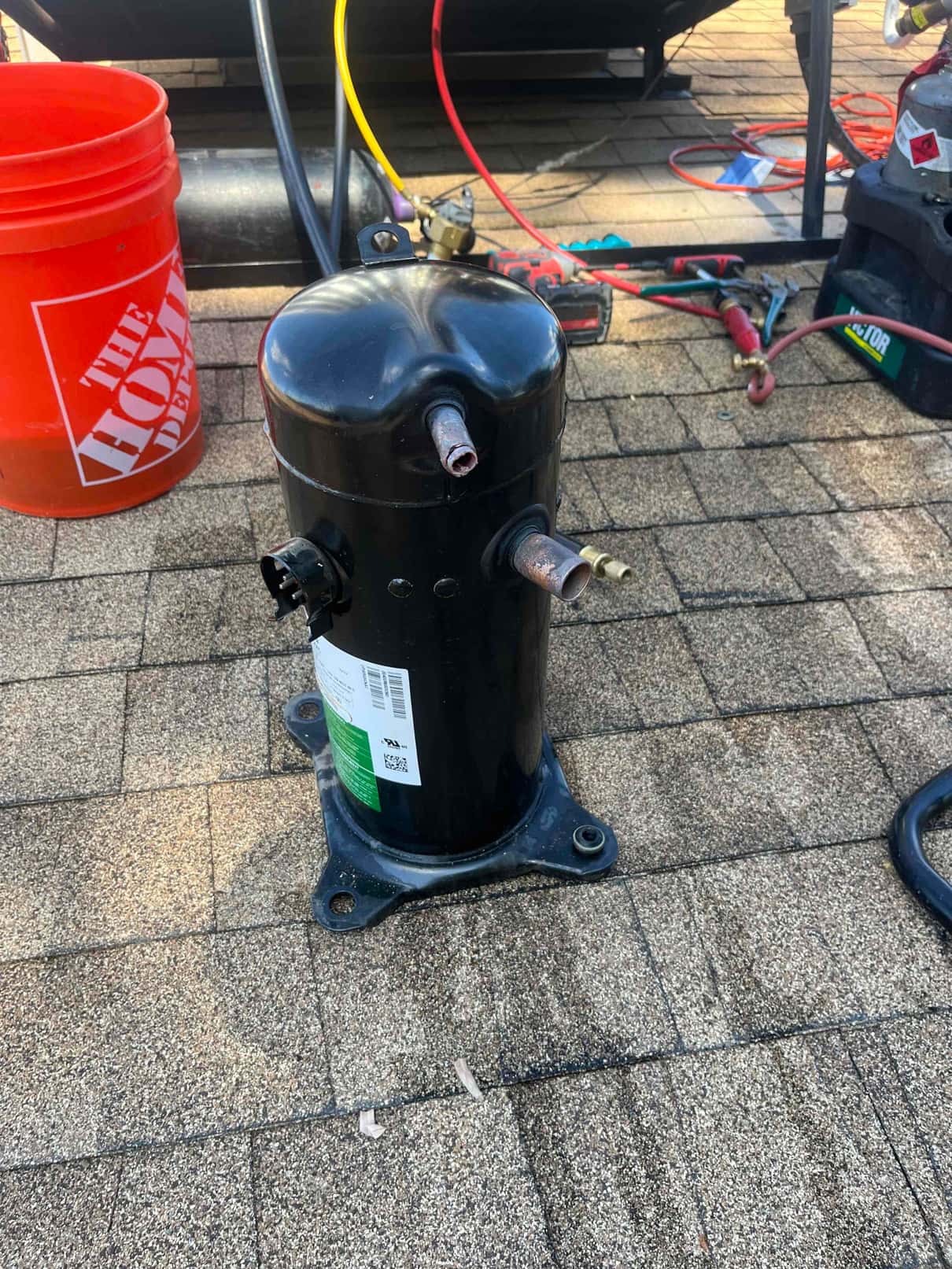 Hermetic refrigeration compressor sits on rooftop shingles with copper suction and discharge stubs exposed and mounting feet visible.