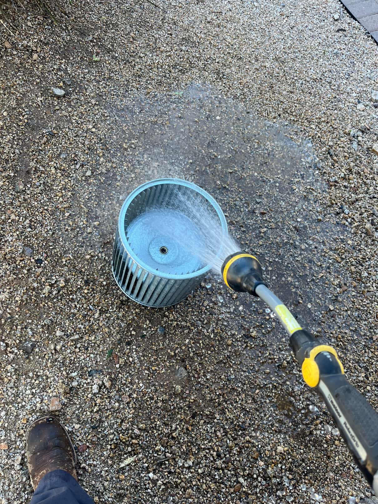 HVAC blower wheel is rinsed with a hose outdoors on gravel.