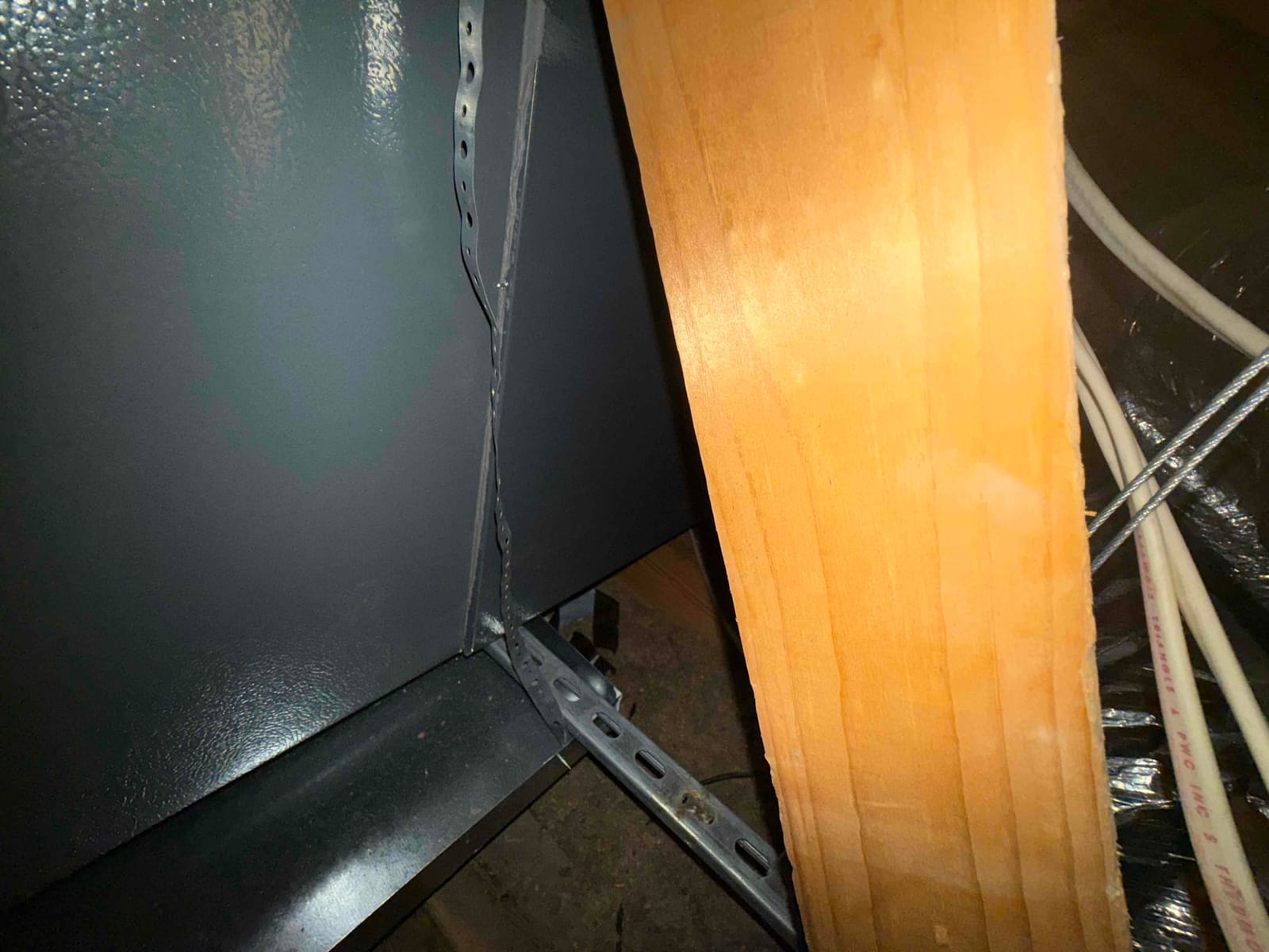 Metal HVAC cabinet in attic with perforated hanger strap and nearby wooden framing, with insulated duct and cables at right.