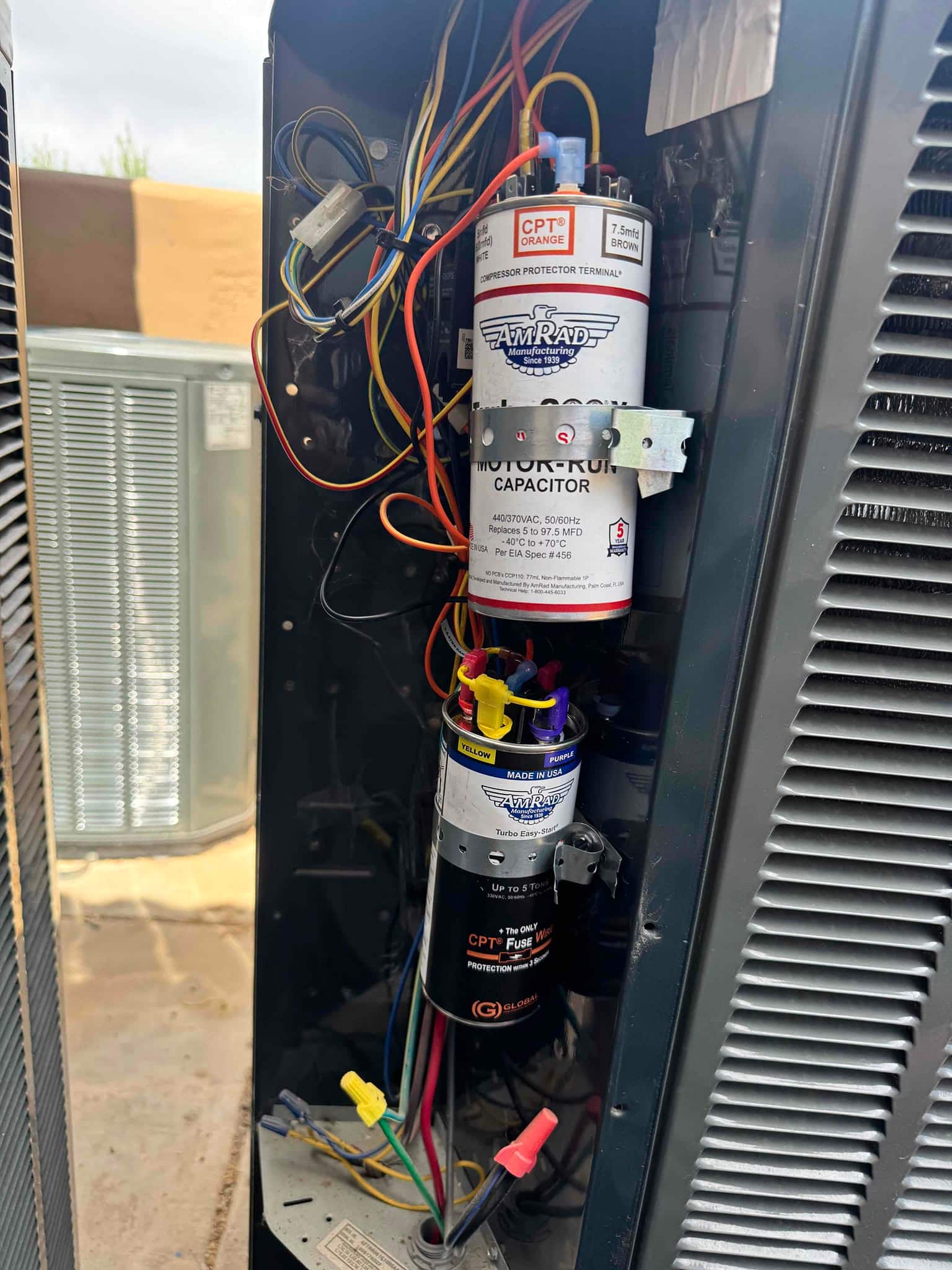 Two motor-run capacitors are mounted in the outdoor condenser’s control compartment, with multiple wires connected and secured.