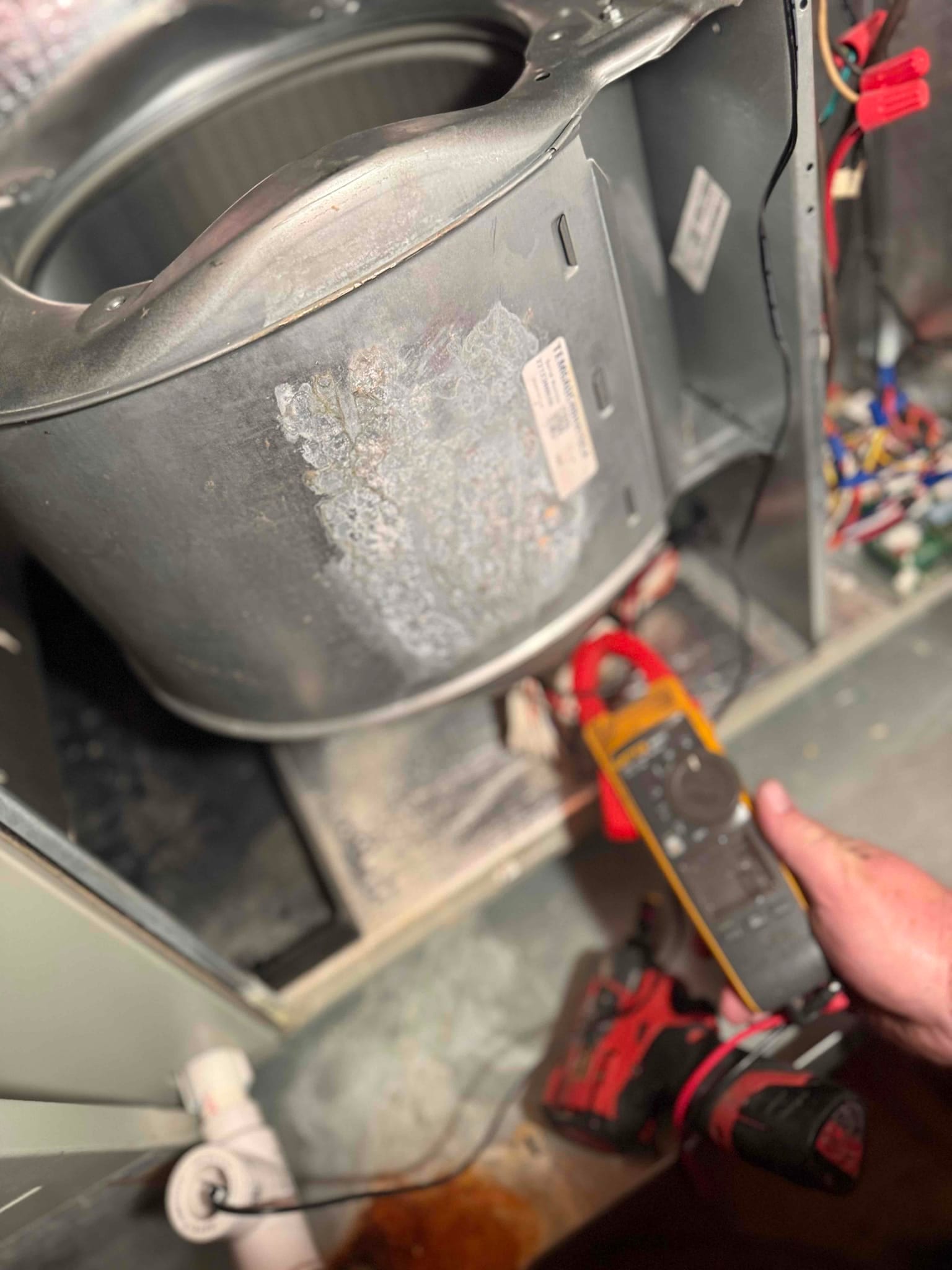 Open air handler cabinet shows metal blower housing and control wiring, with a clamp meter positioned on motor conductors.