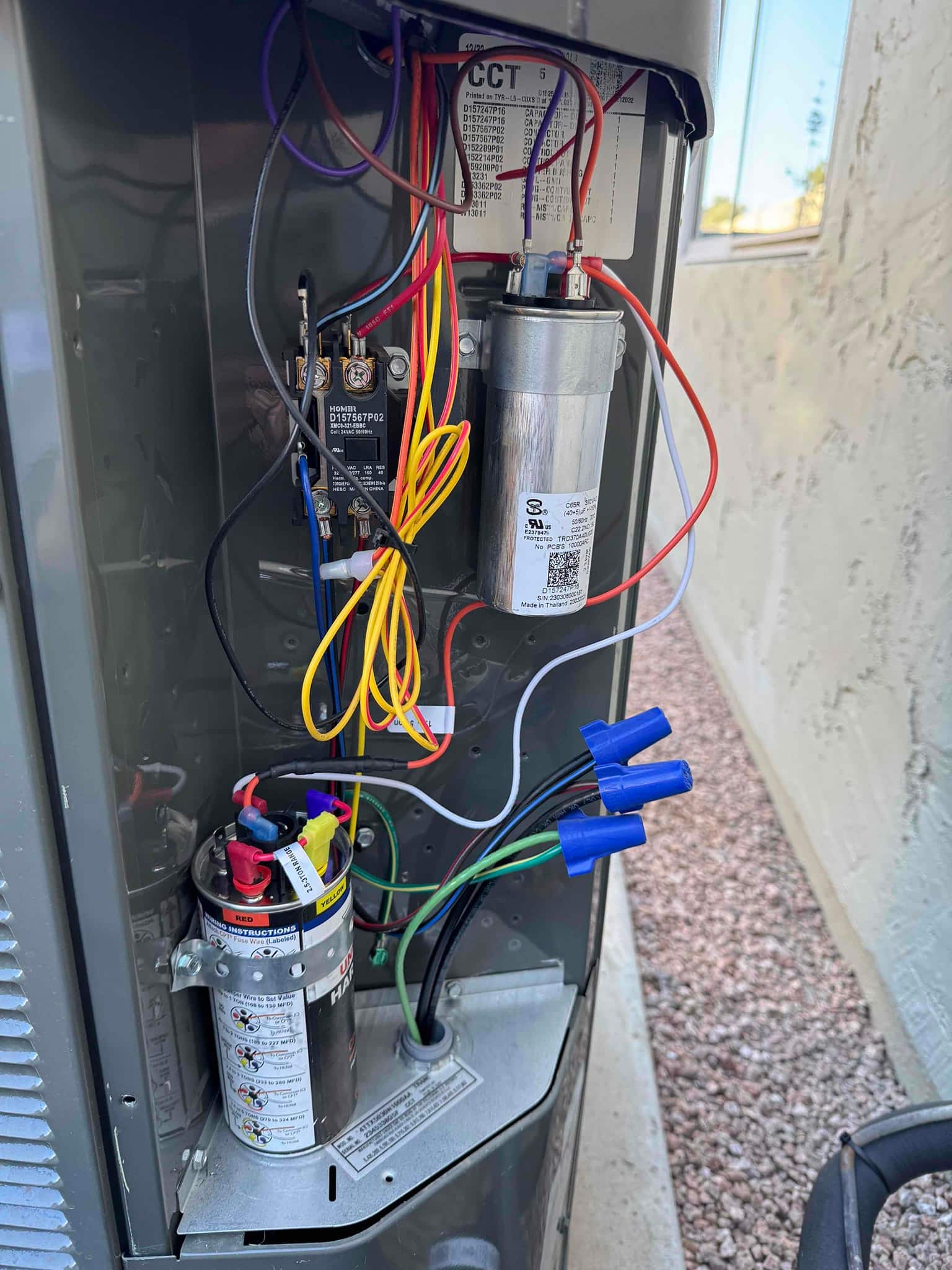 Open outdoor condenser control panel showing dual‑run capacitor, second capacitor, contactor, wiring harnesses, and secured wire‑nut connections.