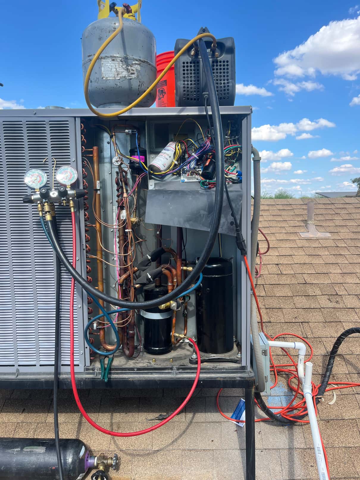 Open rooftop HVAC unit with manifold gauges connected, compressors and copper lines exposed, recovery cylinder and machine positioned on top during service.