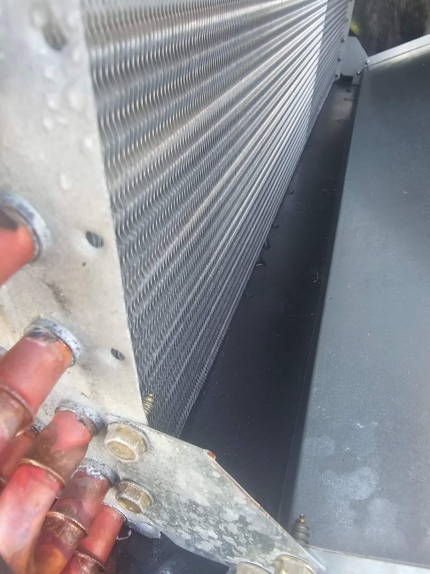 Outdoor condenser coil fins and connected copper refrigerant tubing are visible, with water droplets and light surface oxidation.