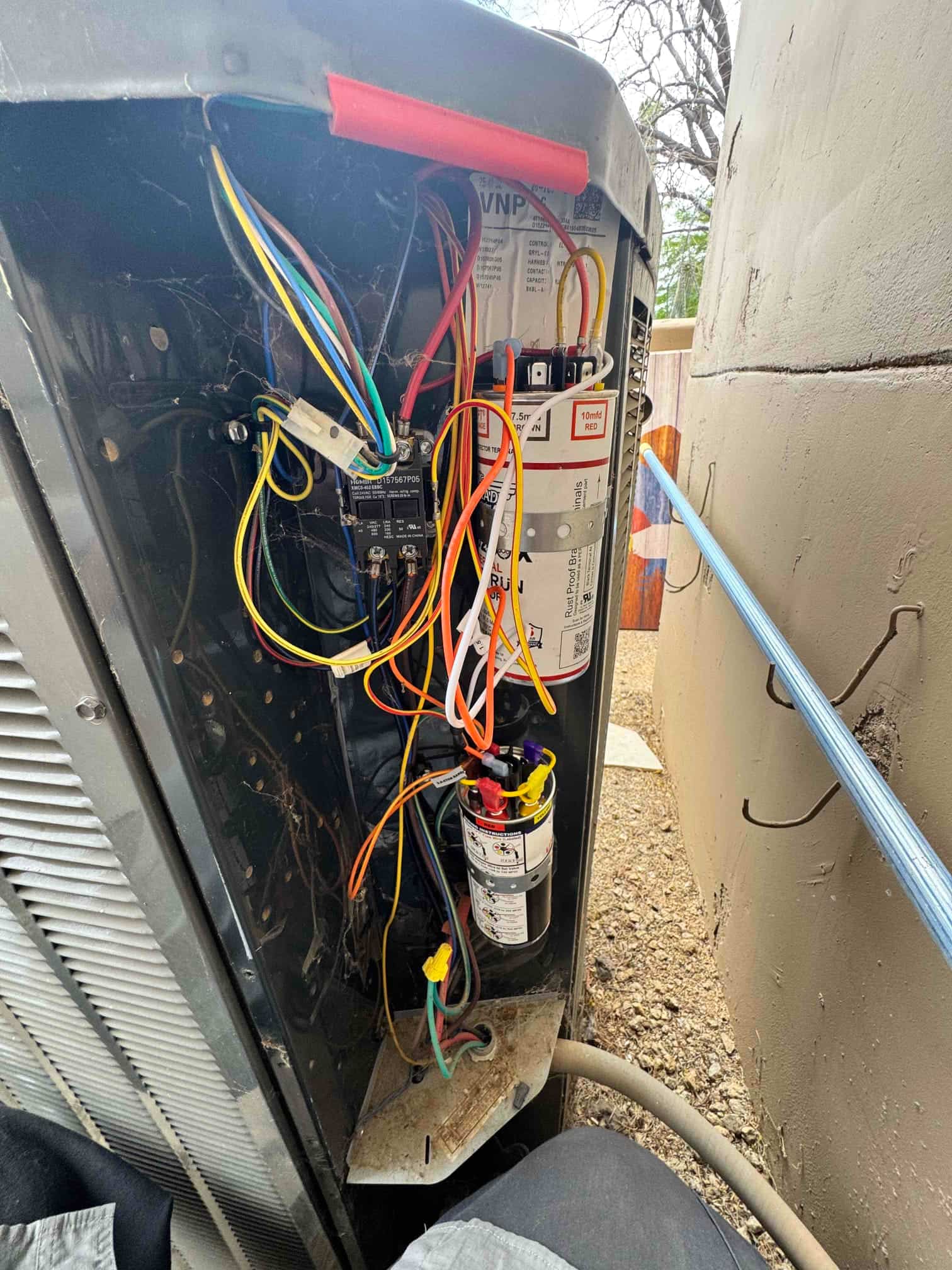 Outdoor condenser control panel open, showing run capacitor, contactor, and connected low-voltage and power wiring inside the cabinet.