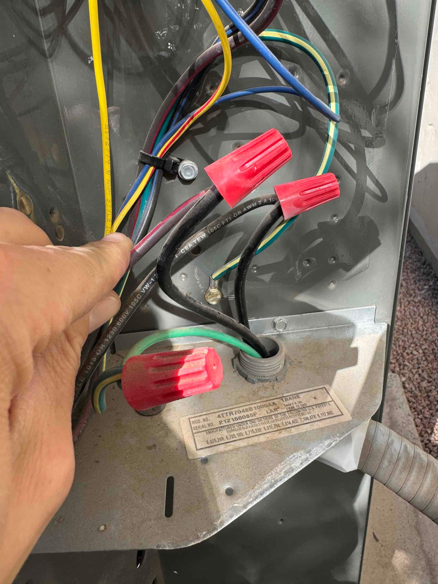Outdoor condenser electrical compartment with multiple conductors and red wire nuts; technician hand holds wires near grounding conductor.