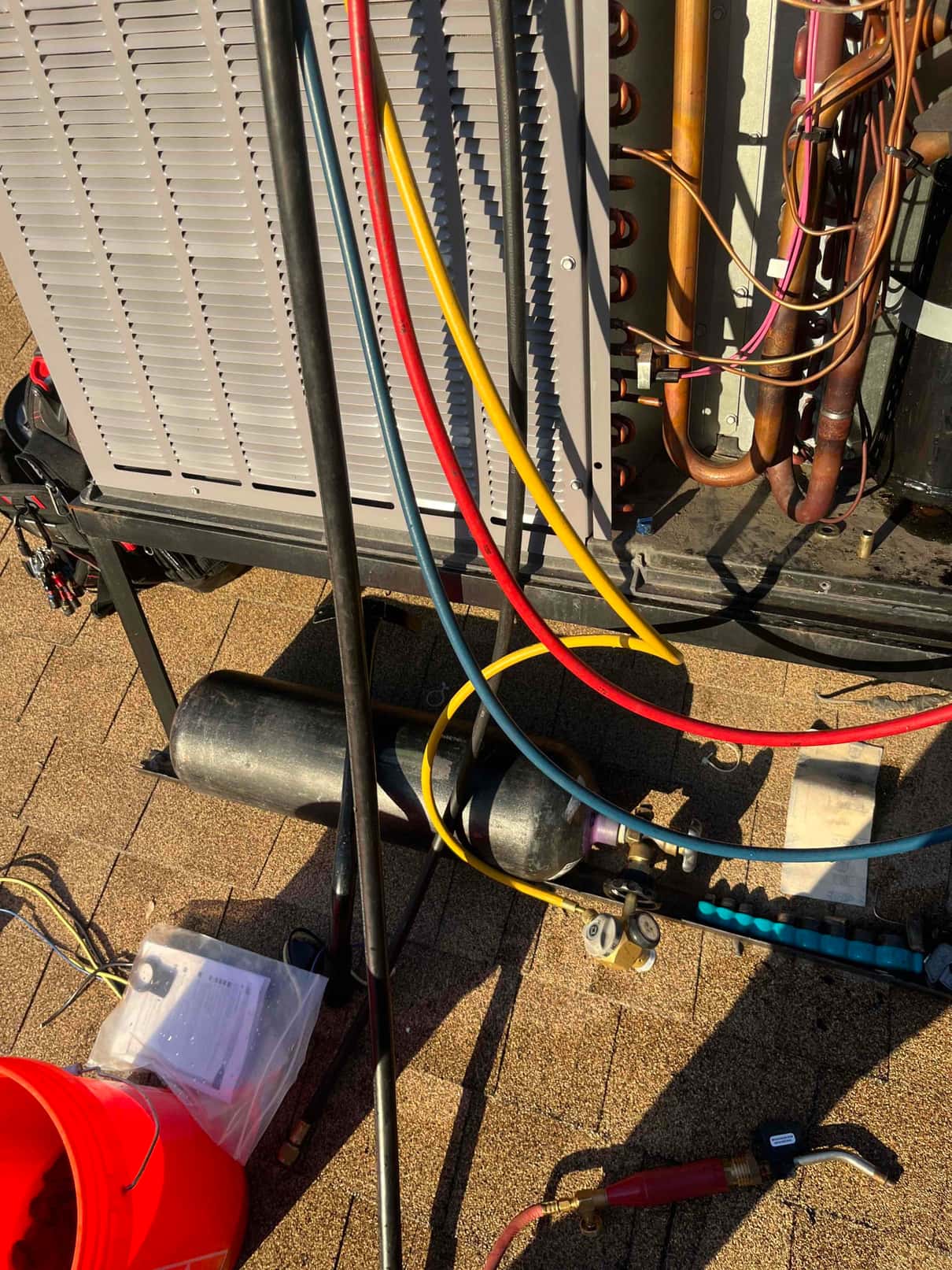 Outdoor condenser with panel open, copper piping exposed, and red, yellow, and blue service hoses routed to a recovery cylinder.