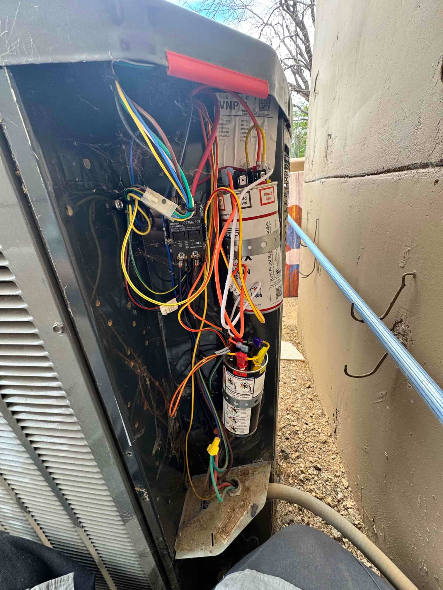 Outdoor condenser service panel open, showing contactor, connected wiring, and a cylindrical capacitor secured inside the unit’s control compartment.
