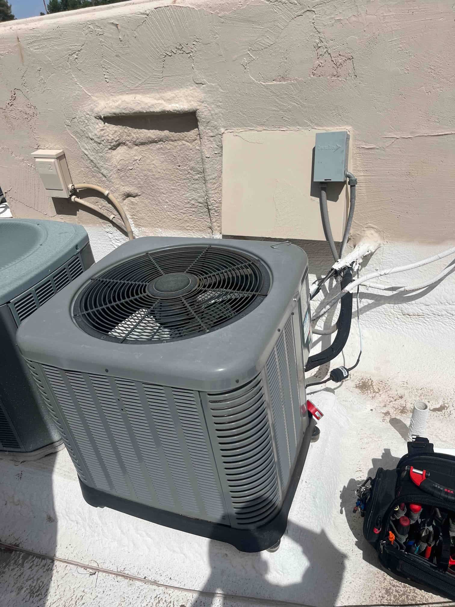 Outdoor condenser unit on rooftop with disconnect box, insulated refrigerant lines, and service valves; technician's tool bag nearby.