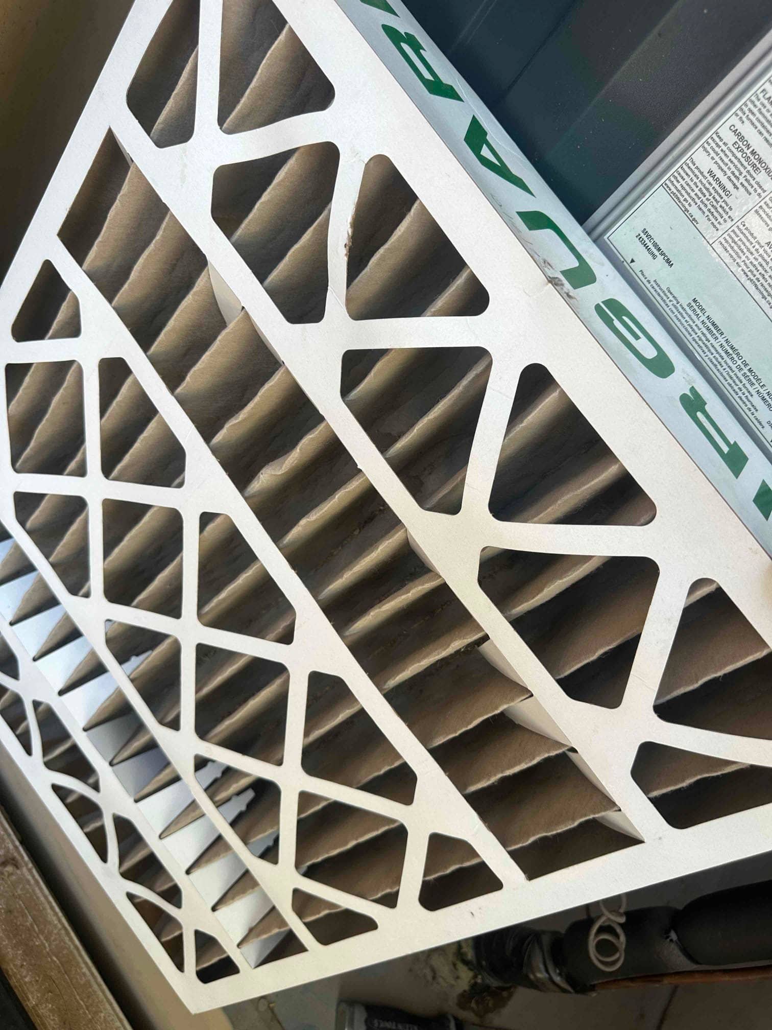 Pleated HVAC air filter with dust on the media resting beside the air handler cabinet.