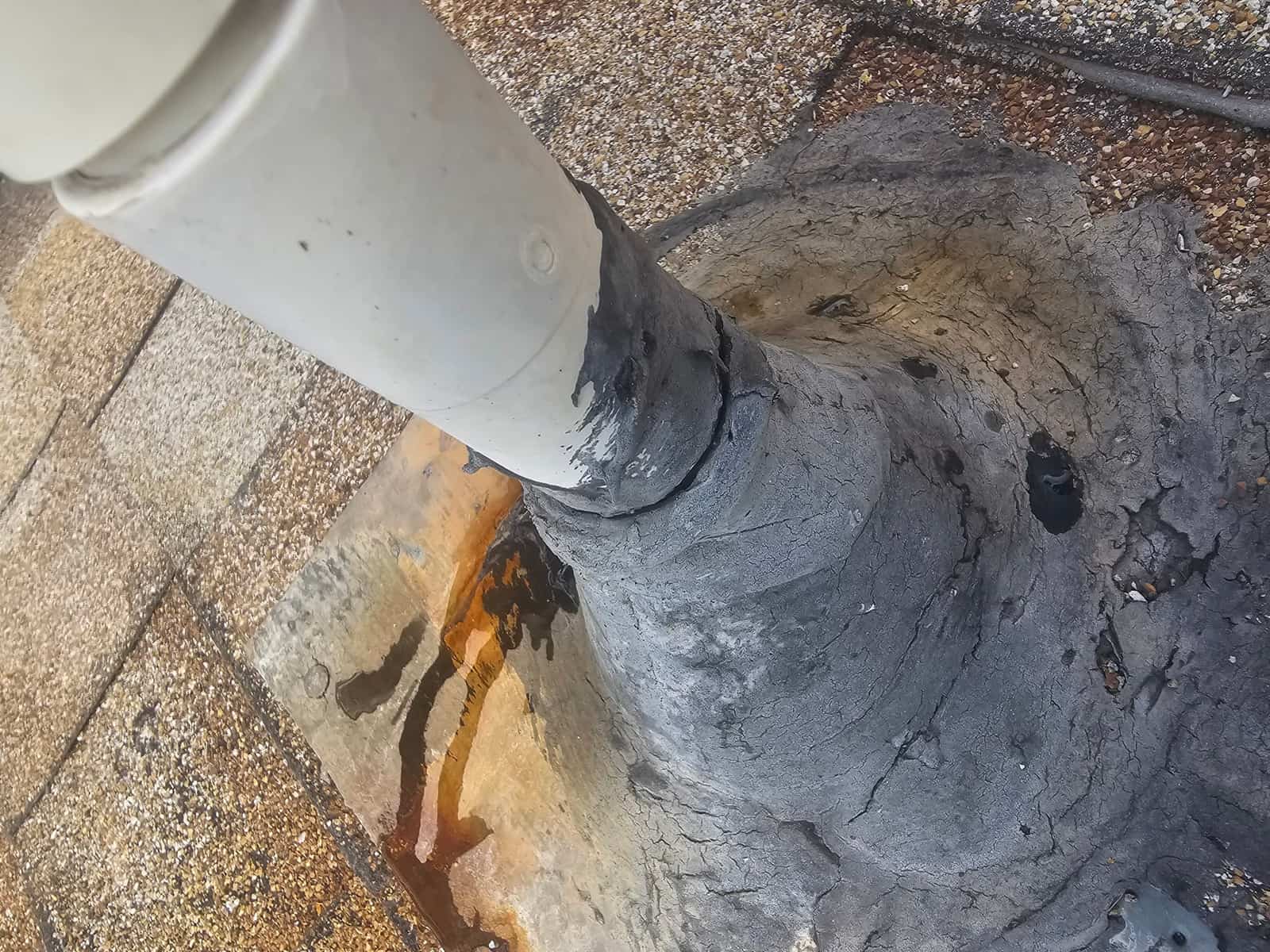 PVC pipe roof penetration shows cracked, weathered sealant and rust-colored staining with water runoff visible nearby.