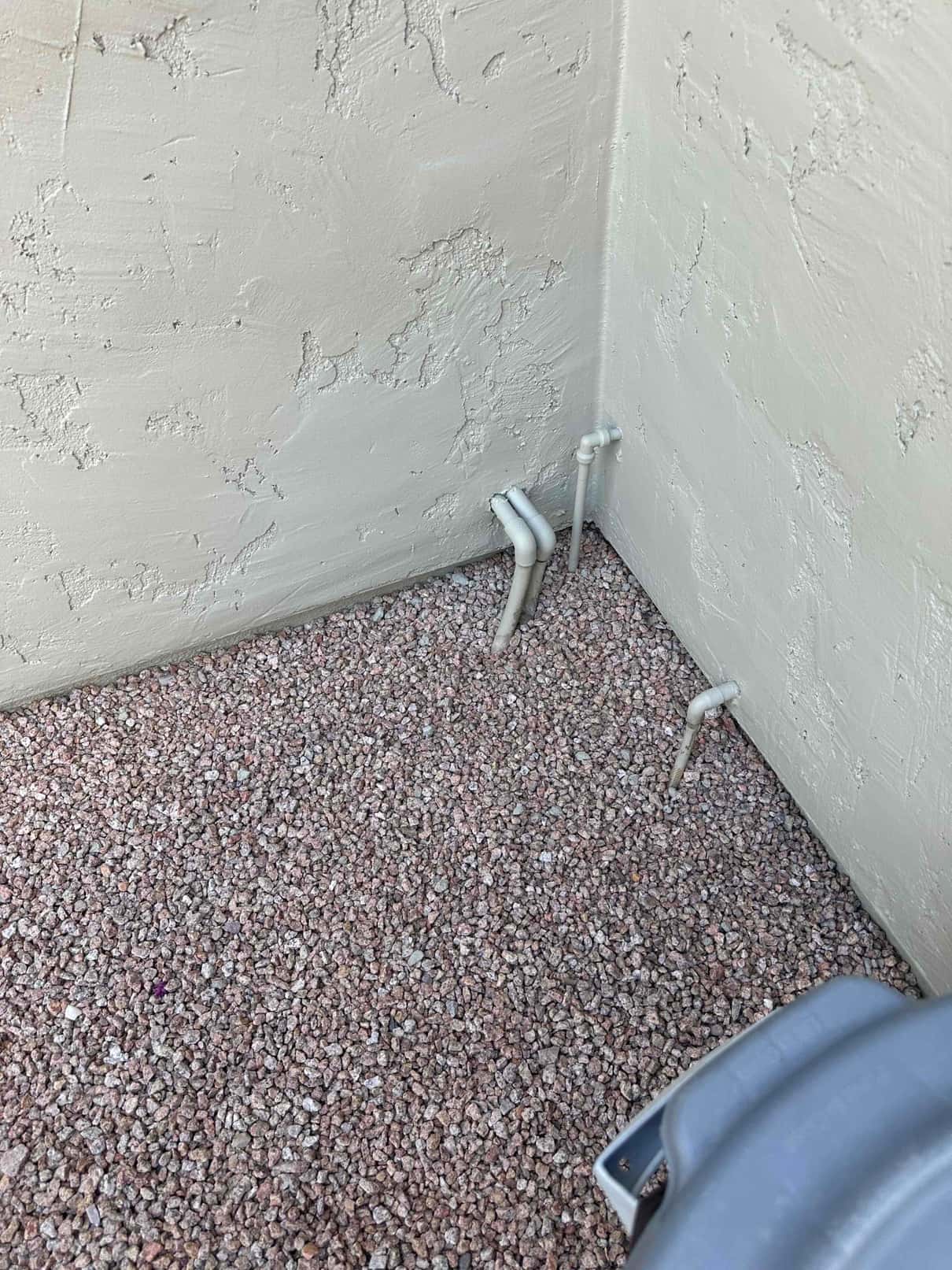 PVC pipes with 90-degree elbows exit the exterior wall near an outdoor HVAC unit on gravel.