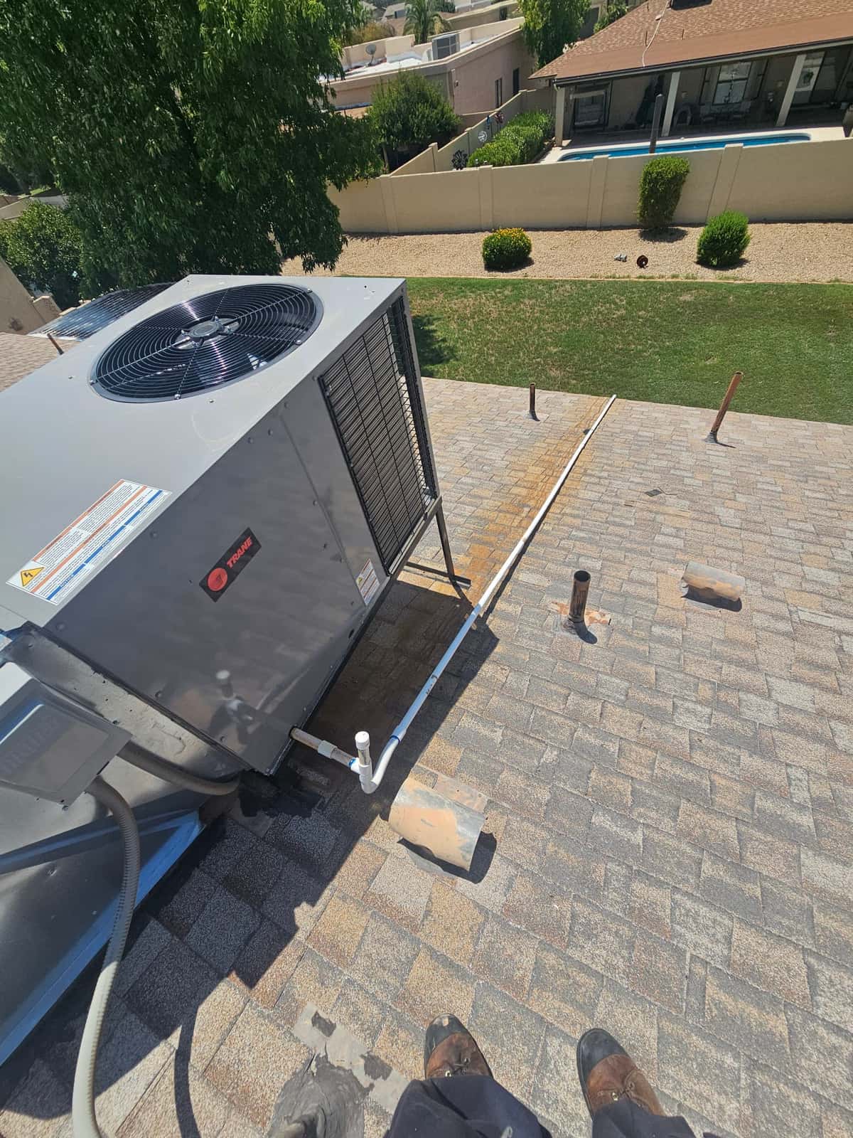 Rooftop packaged HVAC unit on shingle roof, visible condenser fan, side grille, condensate drain piping, and electrical disconnect.