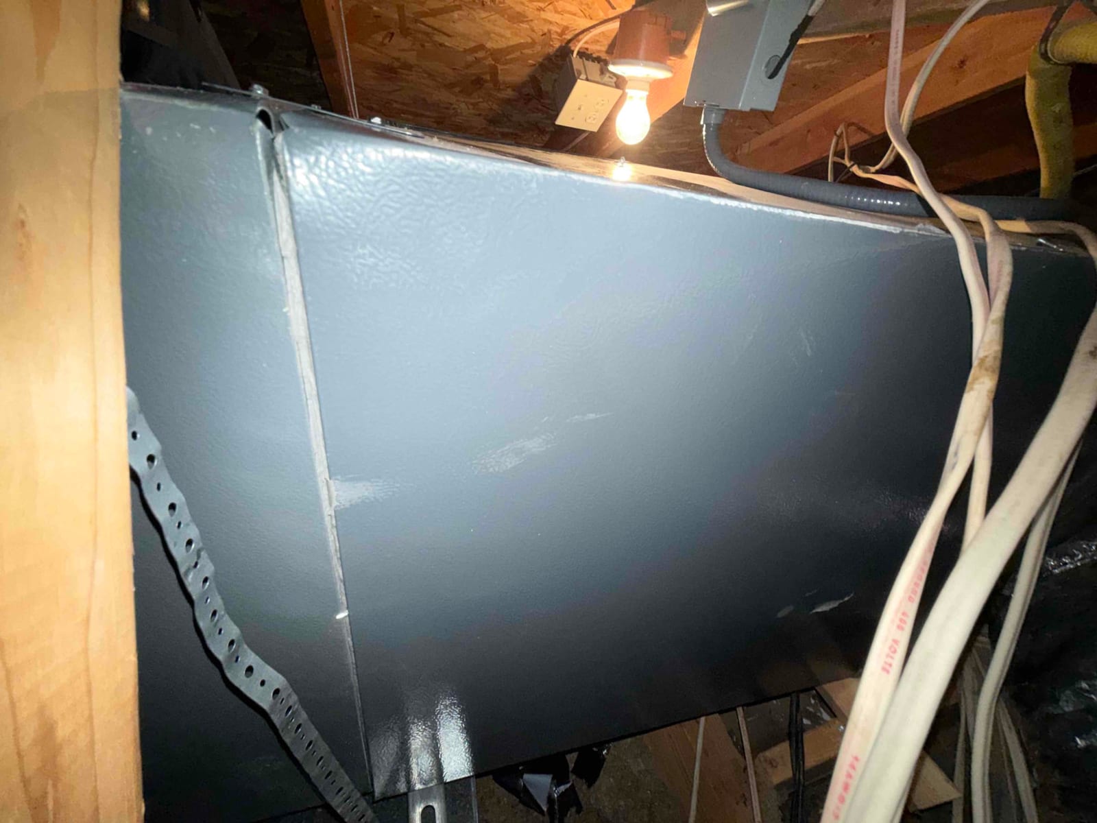 Sheet-metal duct enclosure in attic with hanging strap, visible seam, nearby electrical junction box and light.