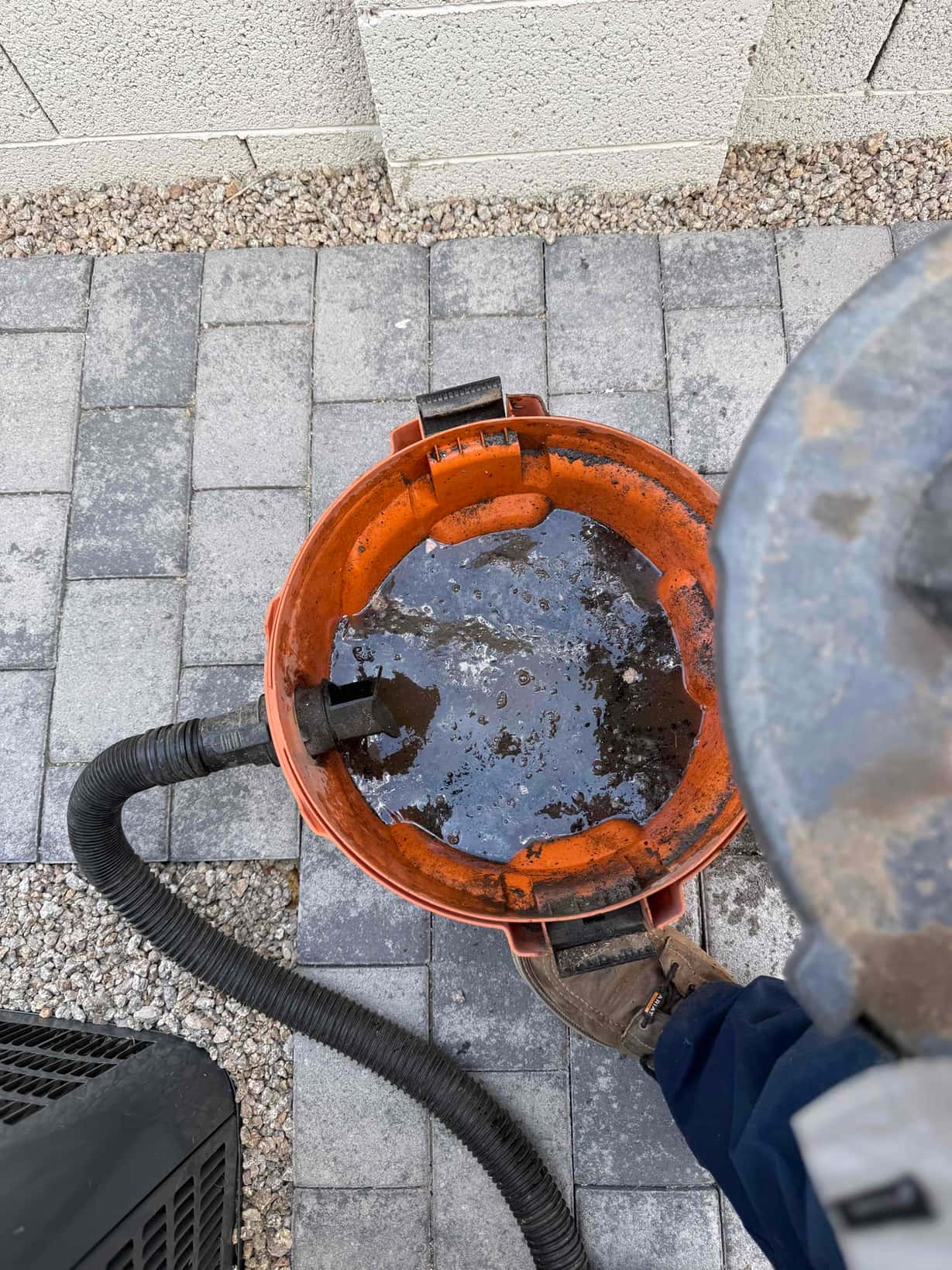 Shop vacuum bucket with dirty water and hose next to outdoor condenser unit on pavers.