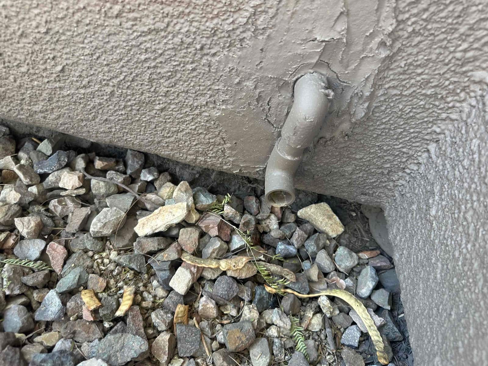 Two short PVC drain outlets exiting the stucco exterior wall above gravel.