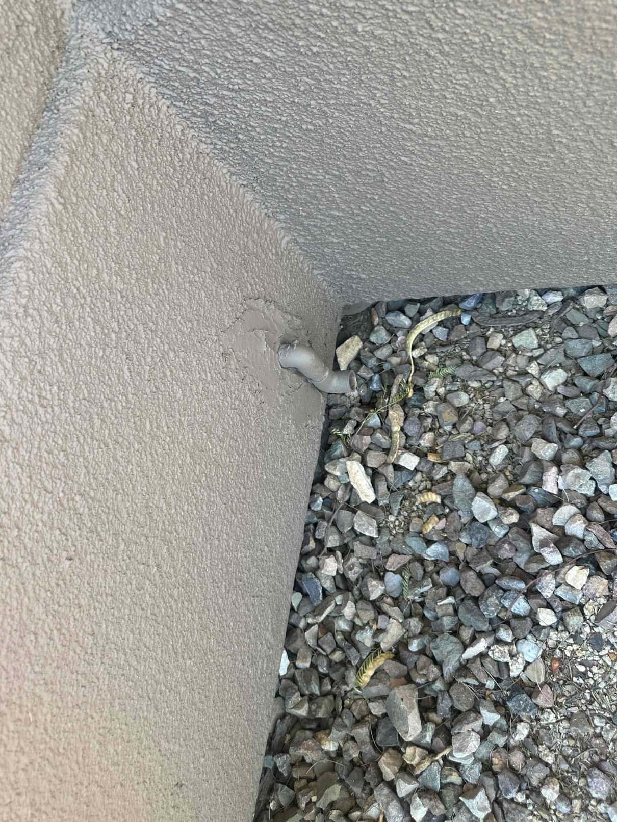 Short PVC drain pipe protrudes from exterior stucco wall above gravel at the foundation.