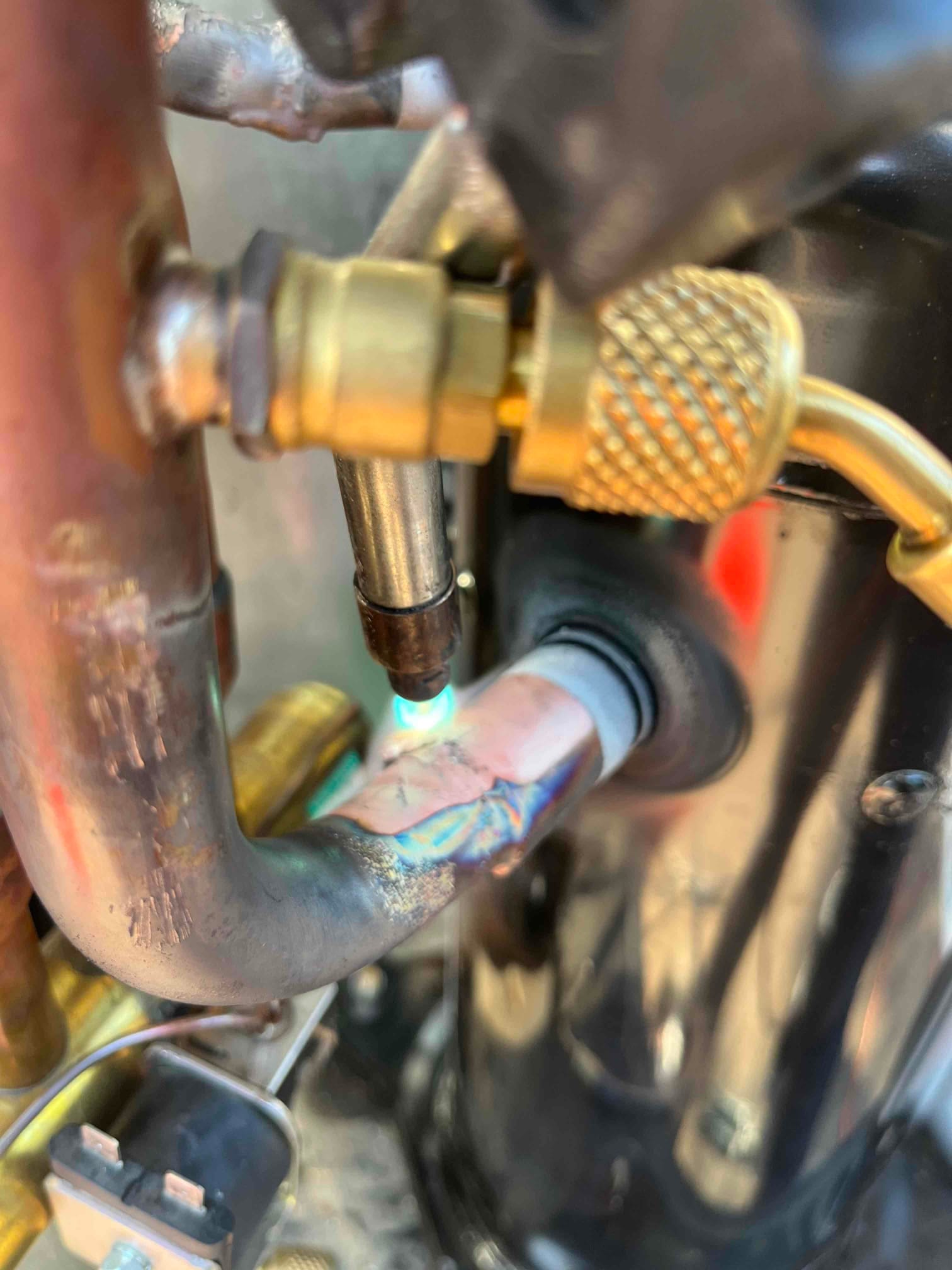 Small torch flame heats a copper refrigerant line joint with visible rainbow discoloration near the compressor and brass service fittings.