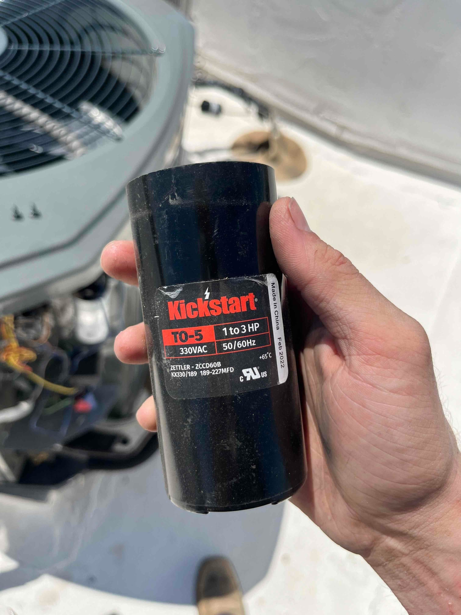 Start capacitor module labeled TO-5 330VAC 1 to 3 HP 50/60Hz, marked 189–227 MFD, held beside outdoor condenser.