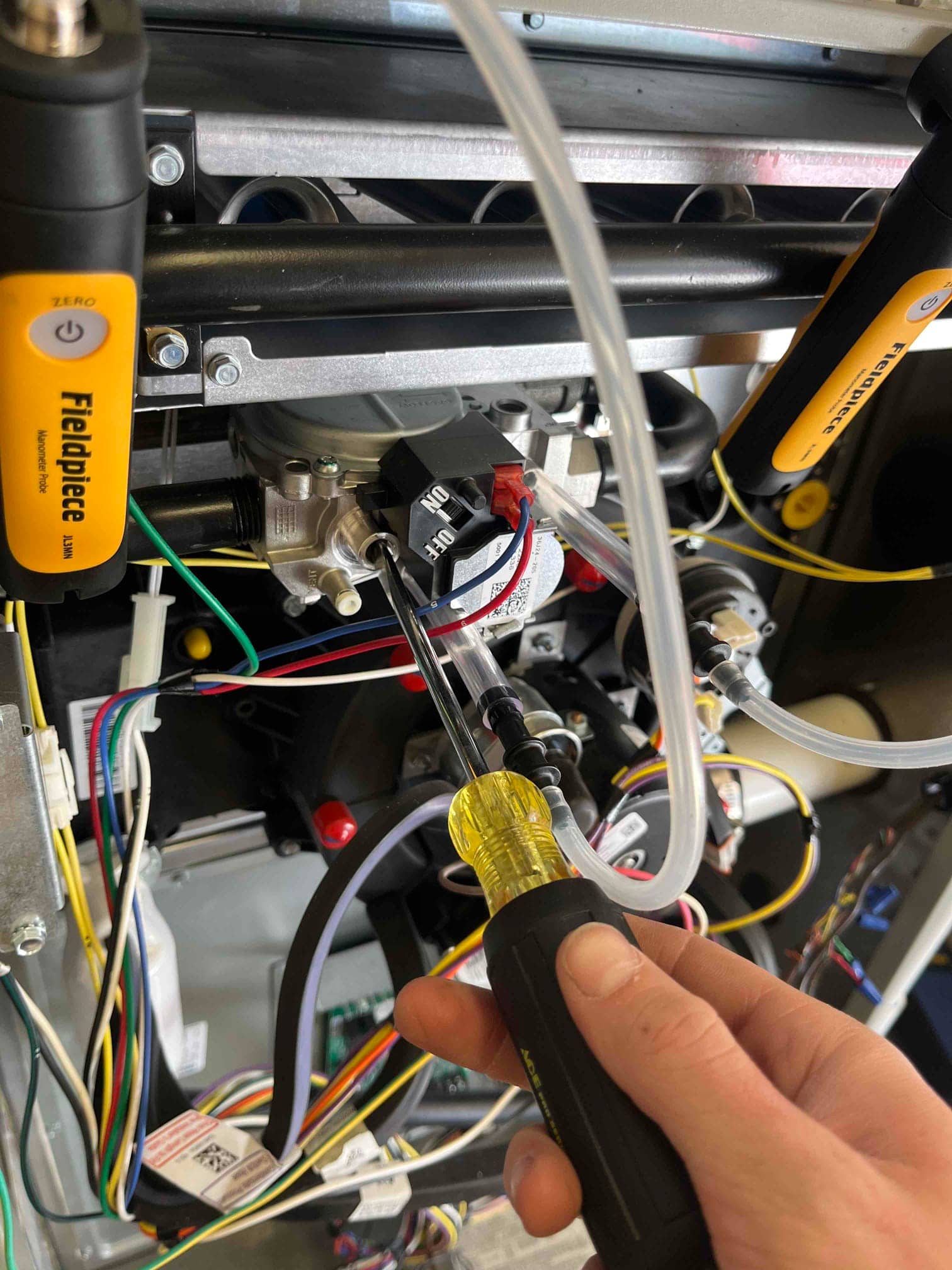 Technician uses a screwdriver at the furnace gas valve while clear tubing and test probes connect to pressure ports.