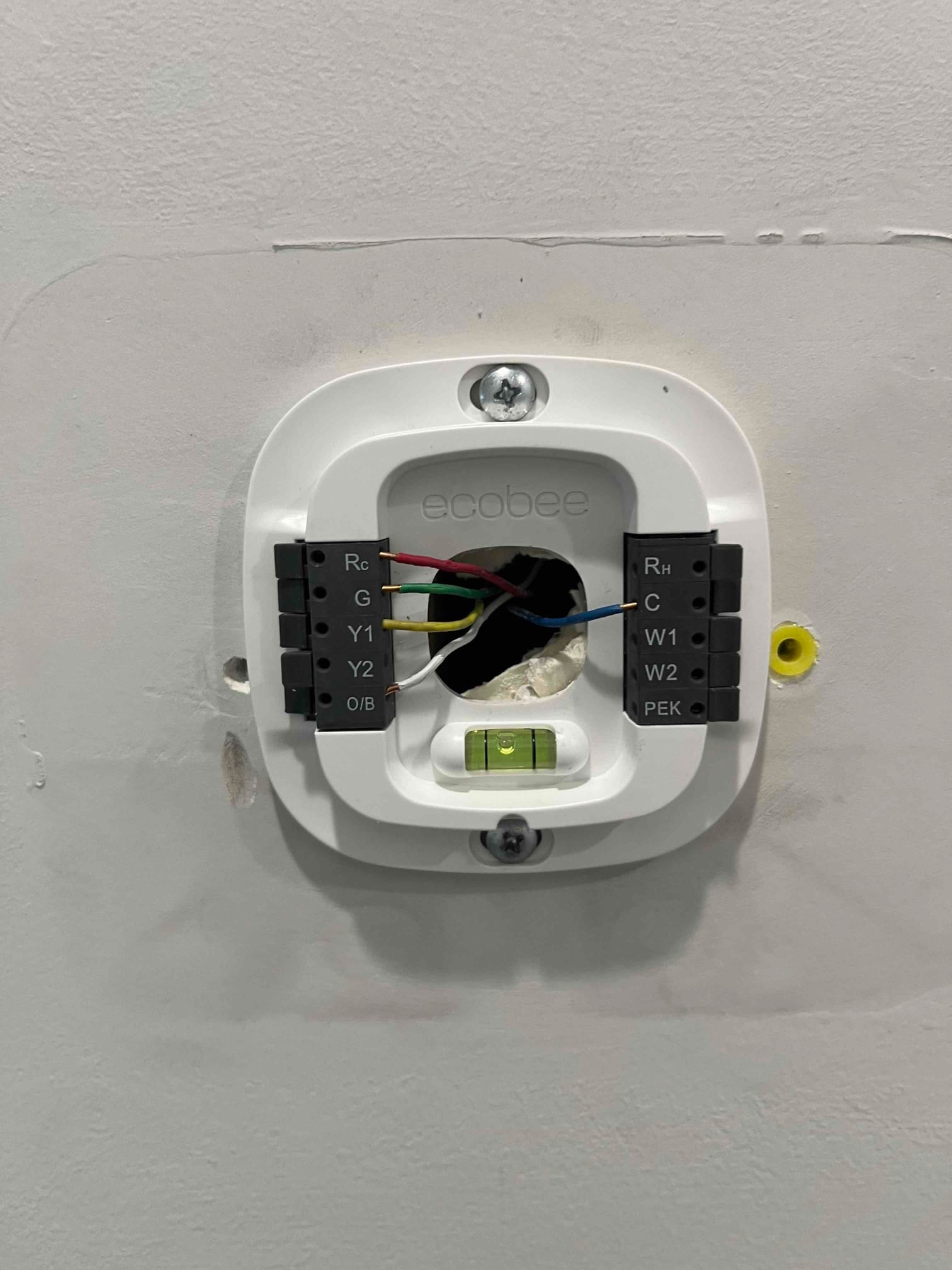 Thermostat wall plate installed on wall, with low-voltage wires connected to labeled terminals and a built-in bubble level.