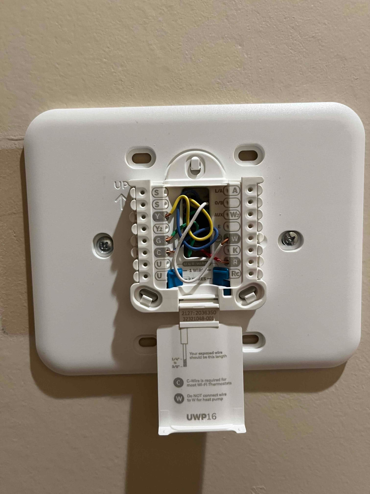 Thermostat wall plate with labeled terminals and multiple low-voltage wires connected, instruction tab open below the mounting base.