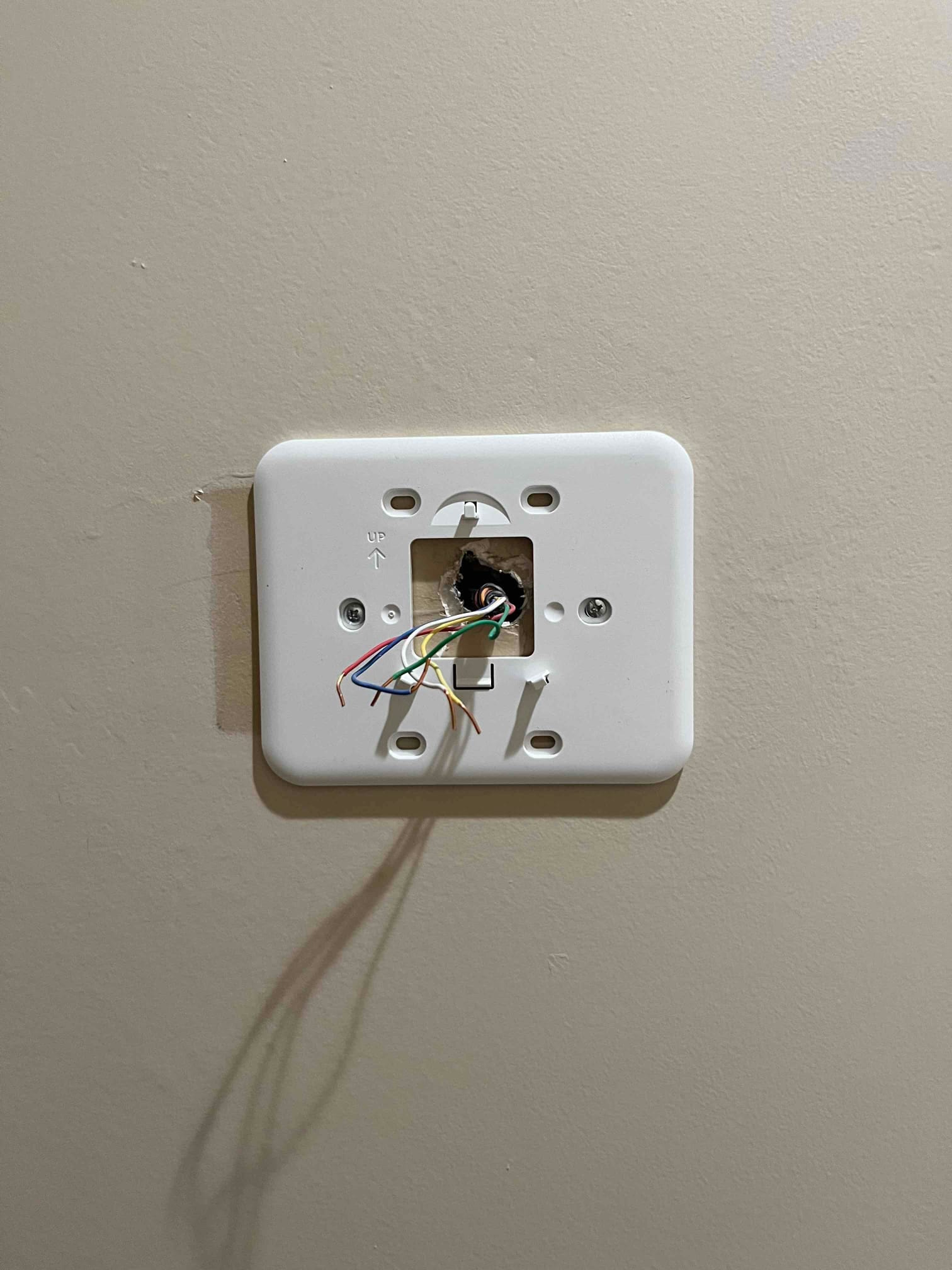 Thermostat wall plate mounted with low-voltage control wires exposed through the center opening on a beige wall.