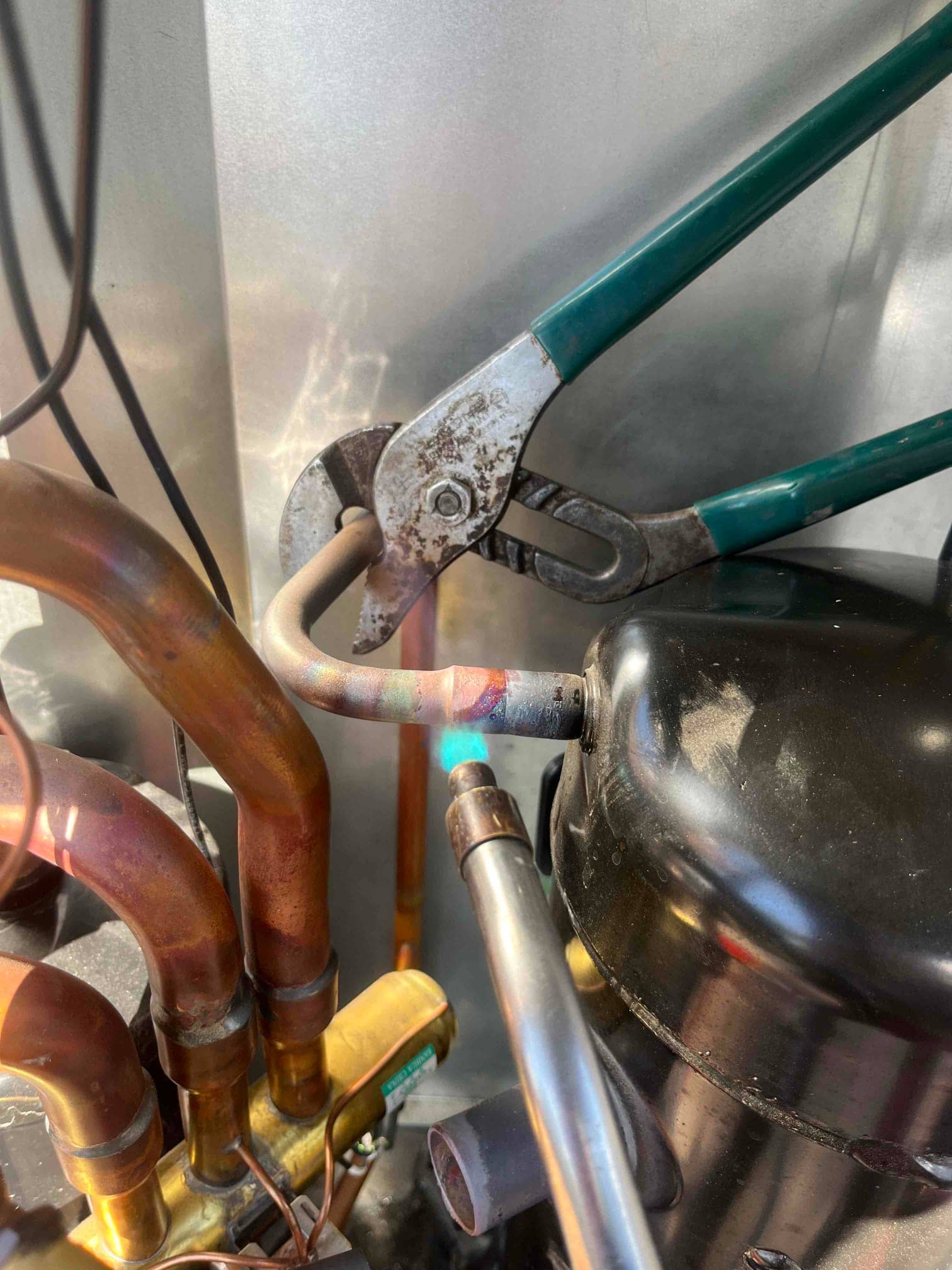Torch heats copper refrigerant line at compressor connection, with pliers holding tubing during brazing.