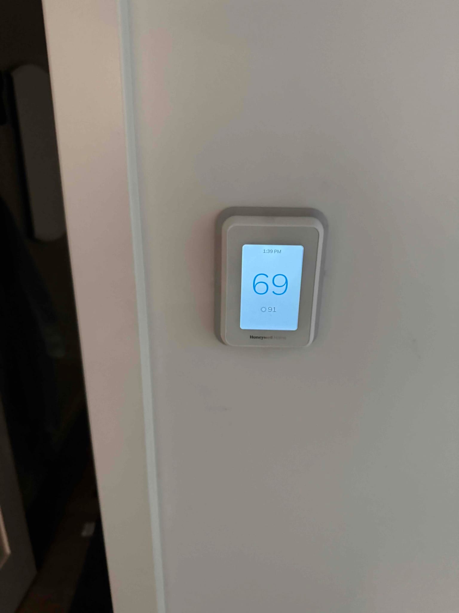 Wall-mounted digital thermostat showing 69 on the screen, installed on an interior wall near a doorway.