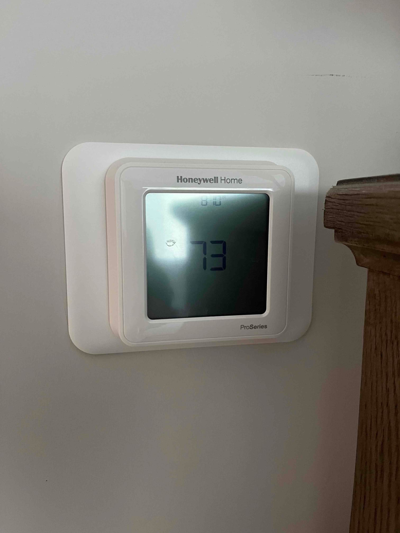 Wall-mounted digital thermostat displays 73 on screen with system icon, installed beside wooden trim.