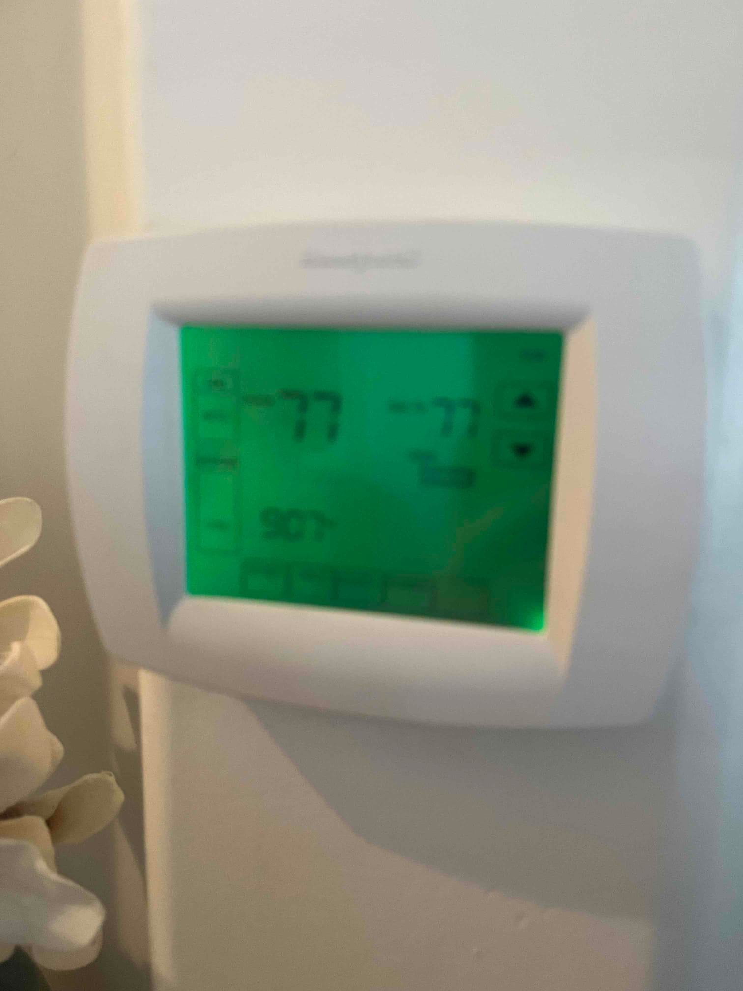 Wall-mounted digital thermostat with green backlight displaying 77 room temperature and 77 setpoint.