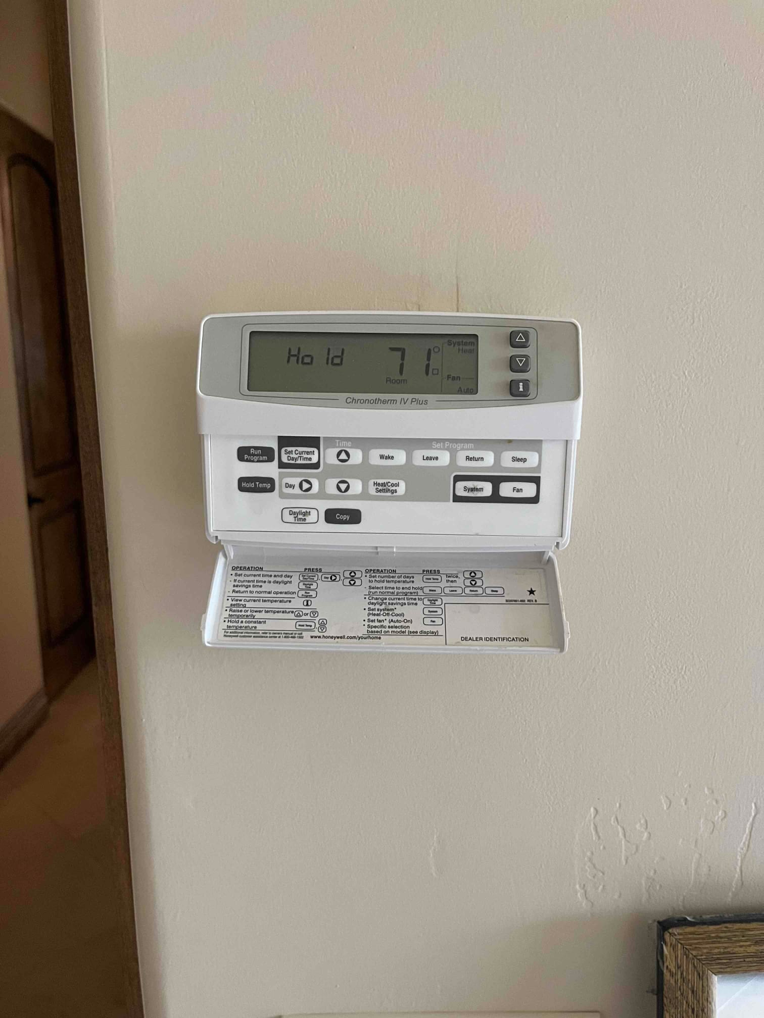 Wall-mounted digital thermostat with programming flap open; display shows Hold, room 71°F, system Heat, and fan Auto.