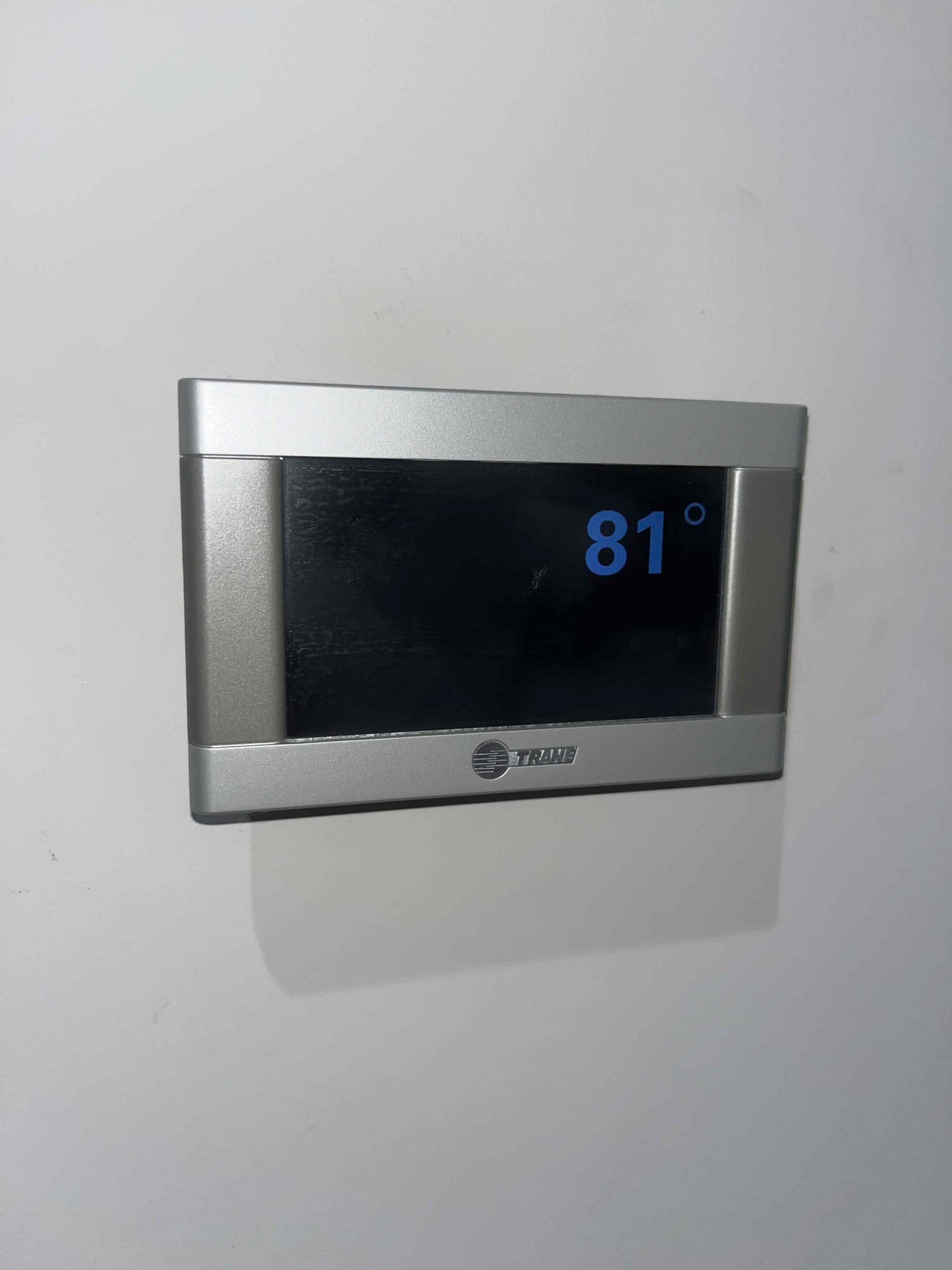 Wall-mounted digital thermostat on a white wall displays 81° on its screen in large blue numerals.