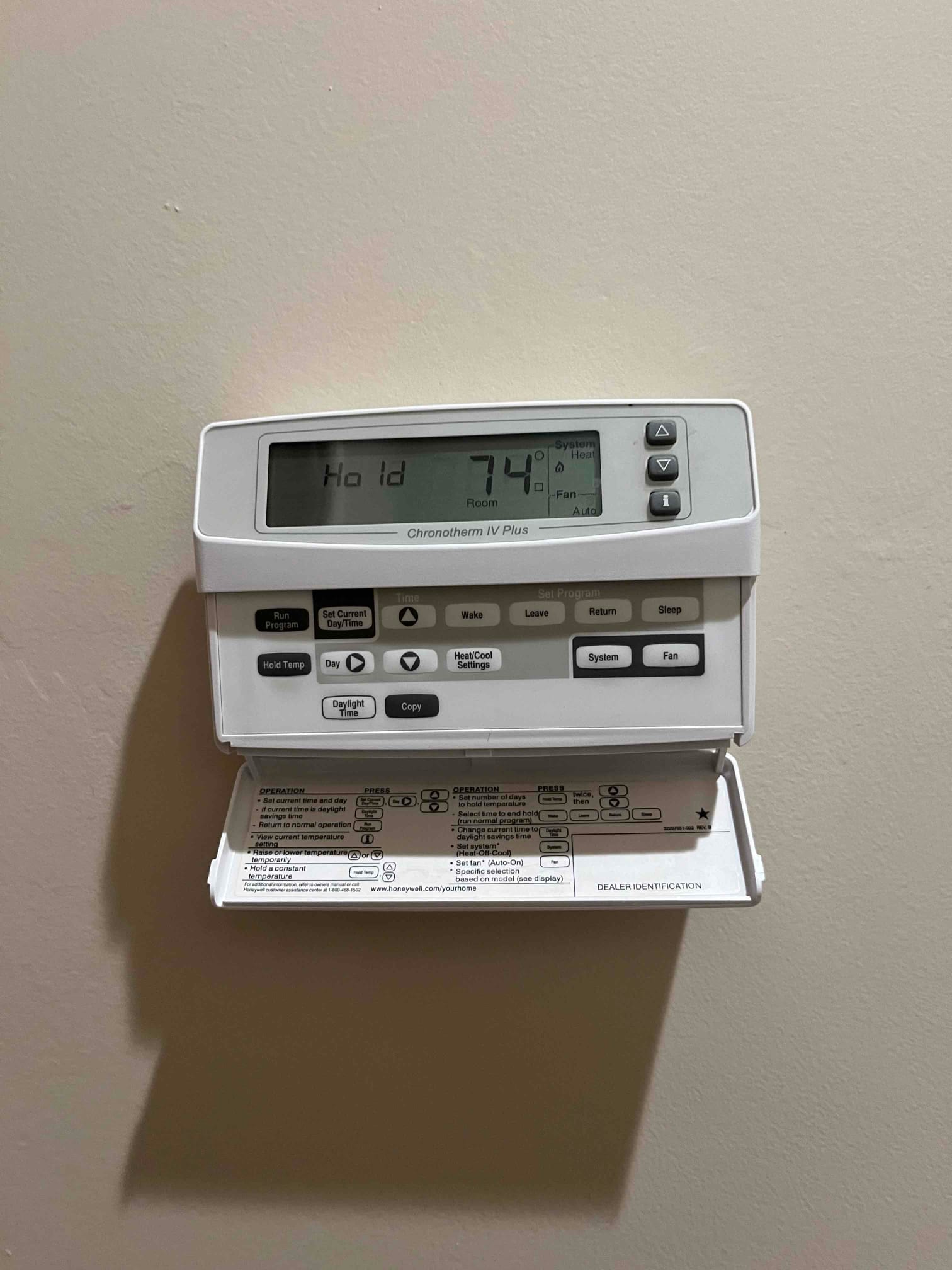 Wall-mounted programmable thermostat with display showing Hold and 74°F room temperature, cover open revealing control instructions.