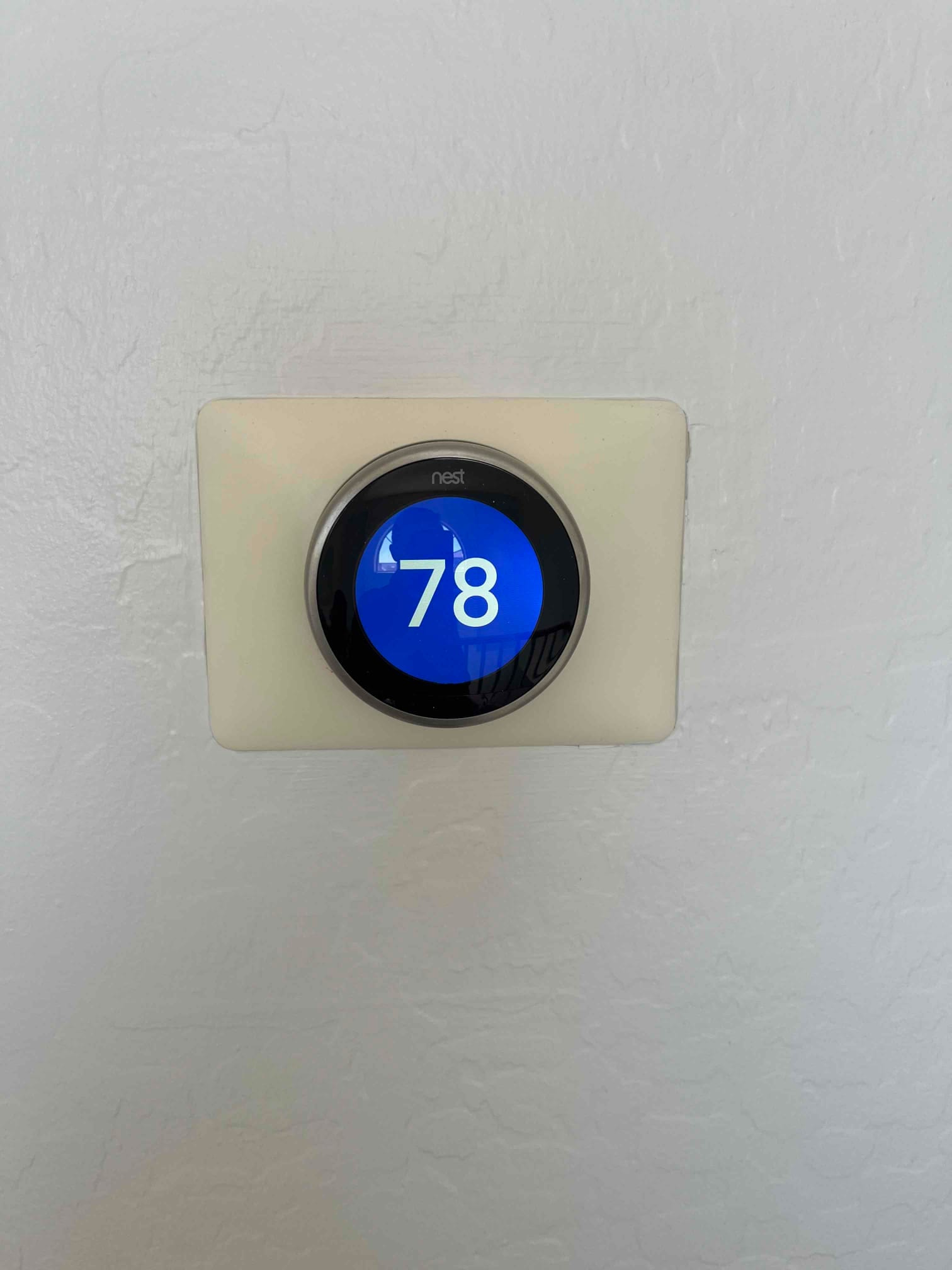 Wall-mounted smart thermostat with circular blue display showing 78 degrees, centered on a rectangular trim plate.