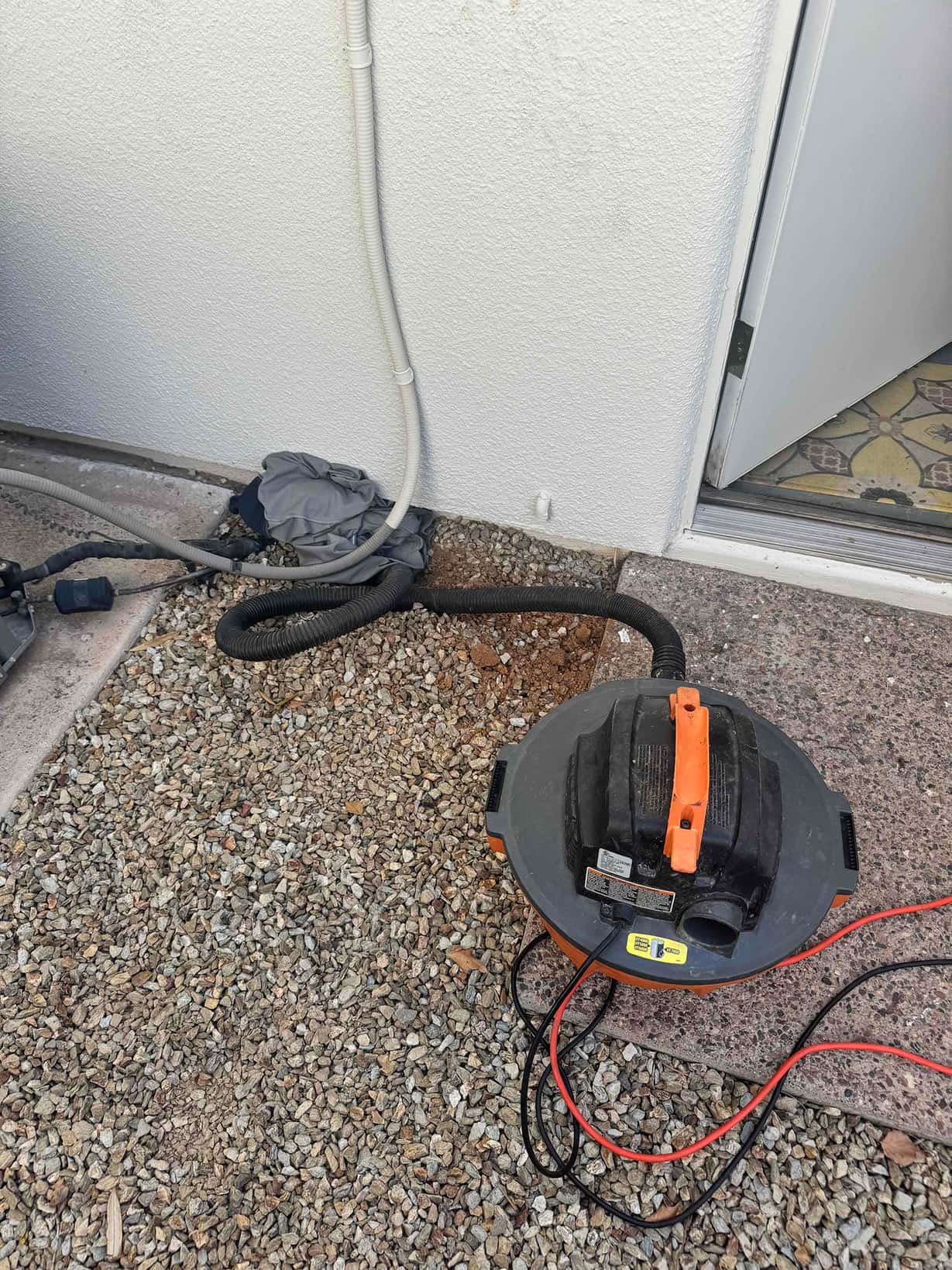 Wet/dry vacuum and flexible hose positioned by exterior wall, next to a small condensate drain outlet near the doorway.