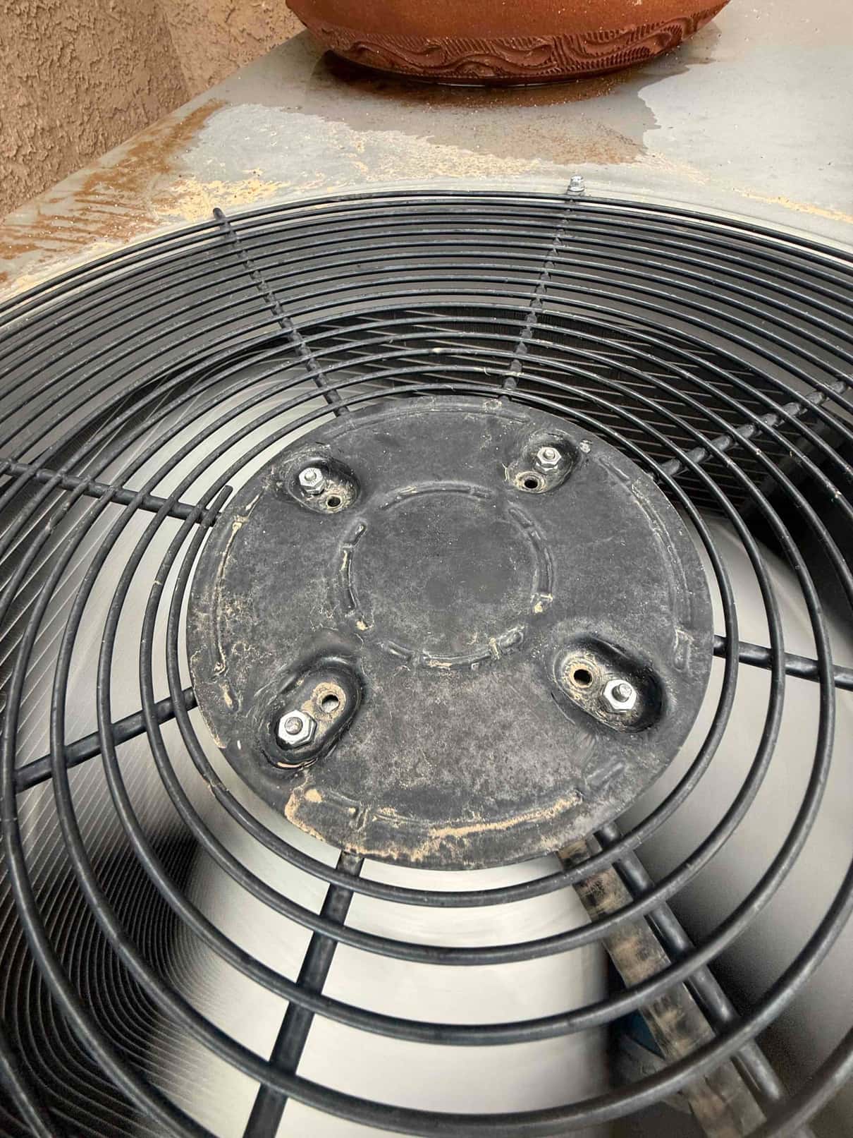Fan guard on an outdoor condenser unit with central mounting plate and hardware exposed.