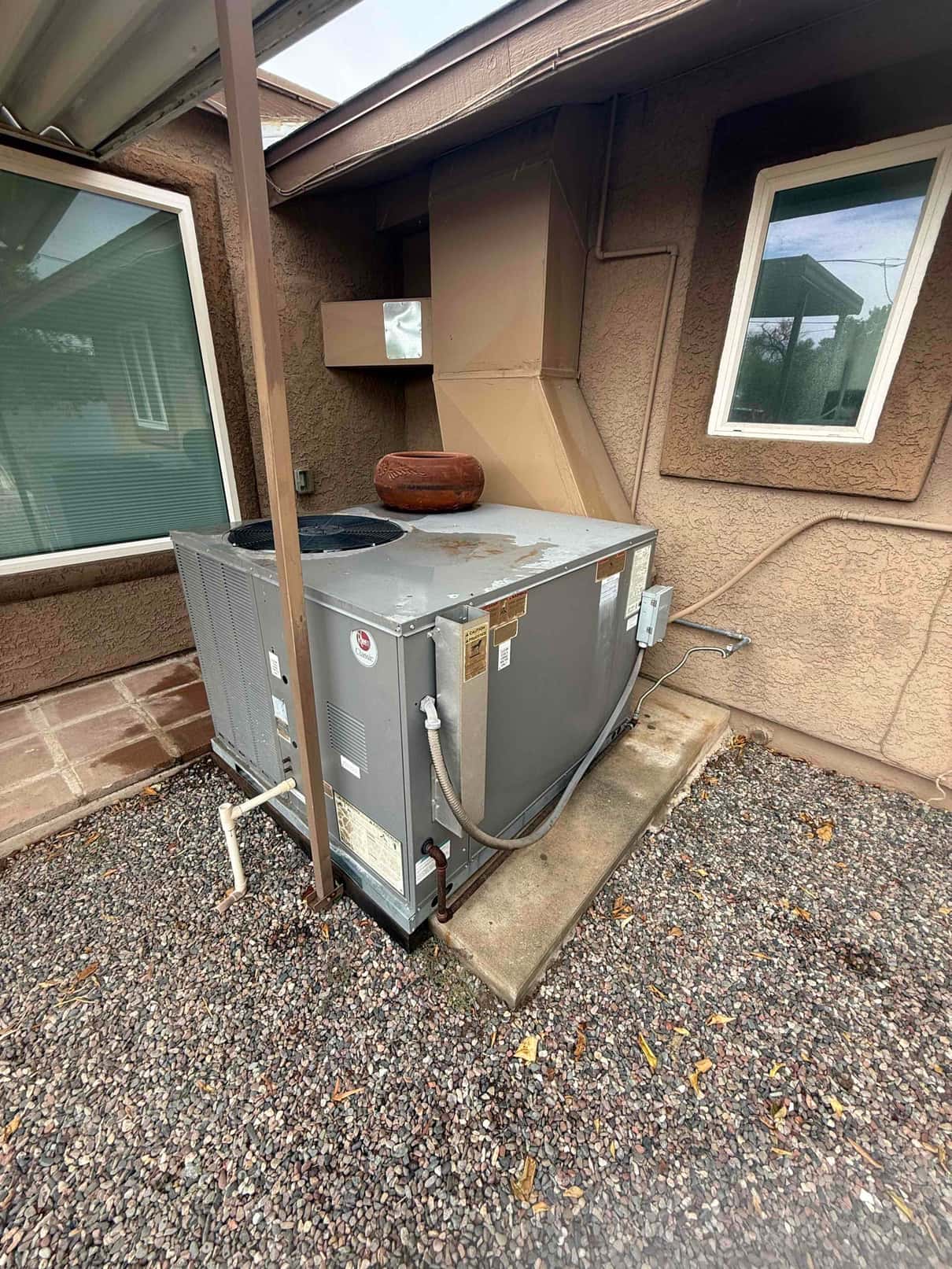Packaged rooftop-style unit at ground level with sheet-metal duct transition, condensate drain, service disconnect, and a rusty pulley resting on top.