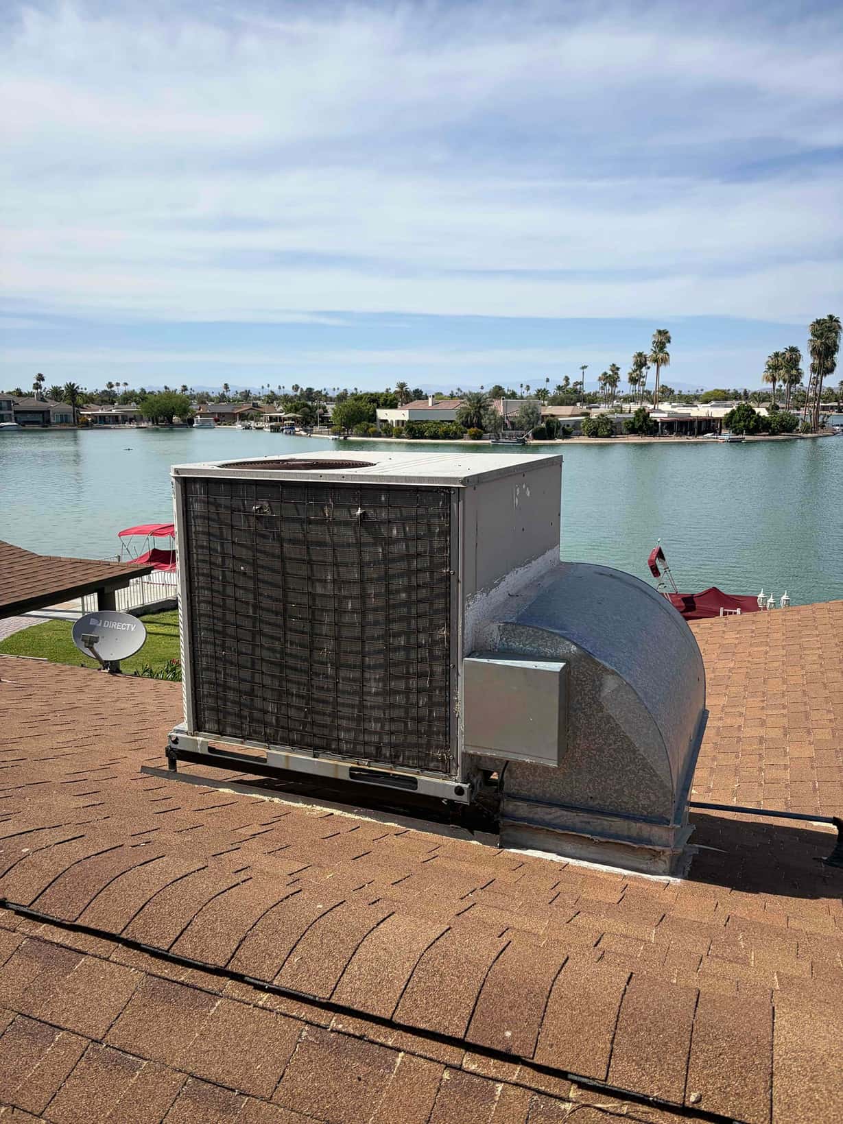 Rooftop HVAC package unit with exposed condenser coil and sheet-metal supply duct on shingle roof.