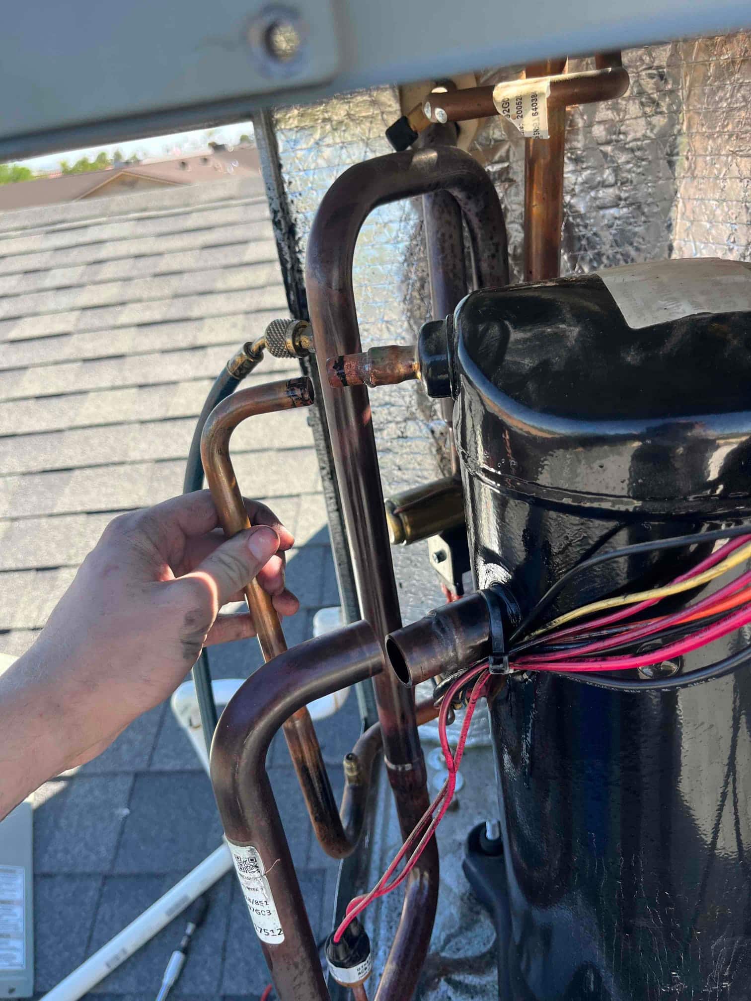 Compressor with wiring harness visible beside open copper refrigerant lines; technician hand holds a tubing section near the connection.