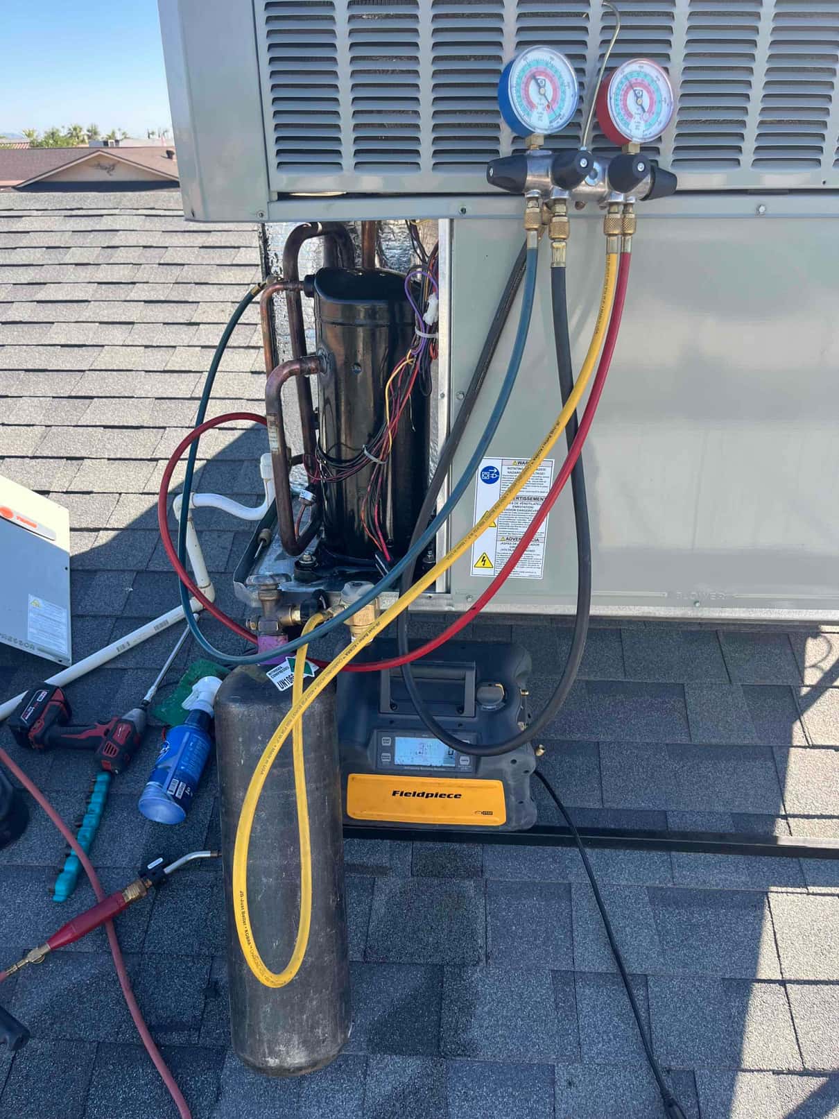 Rooftop package unit with access panel open, compressor exposed, manifold gauges connected, and a refrigerant cylinder with a digital machine set on the shingles.