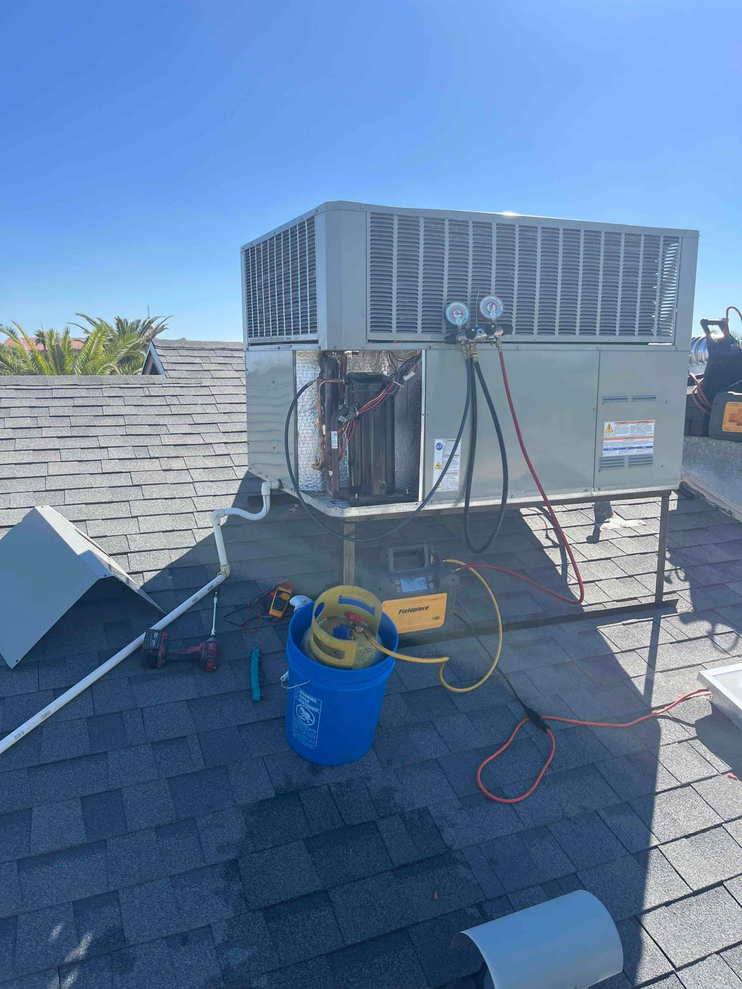 Rooftop package unit with service panel open, manifold gauges connected, recovery cylinder and recovery machine staged on the roof.