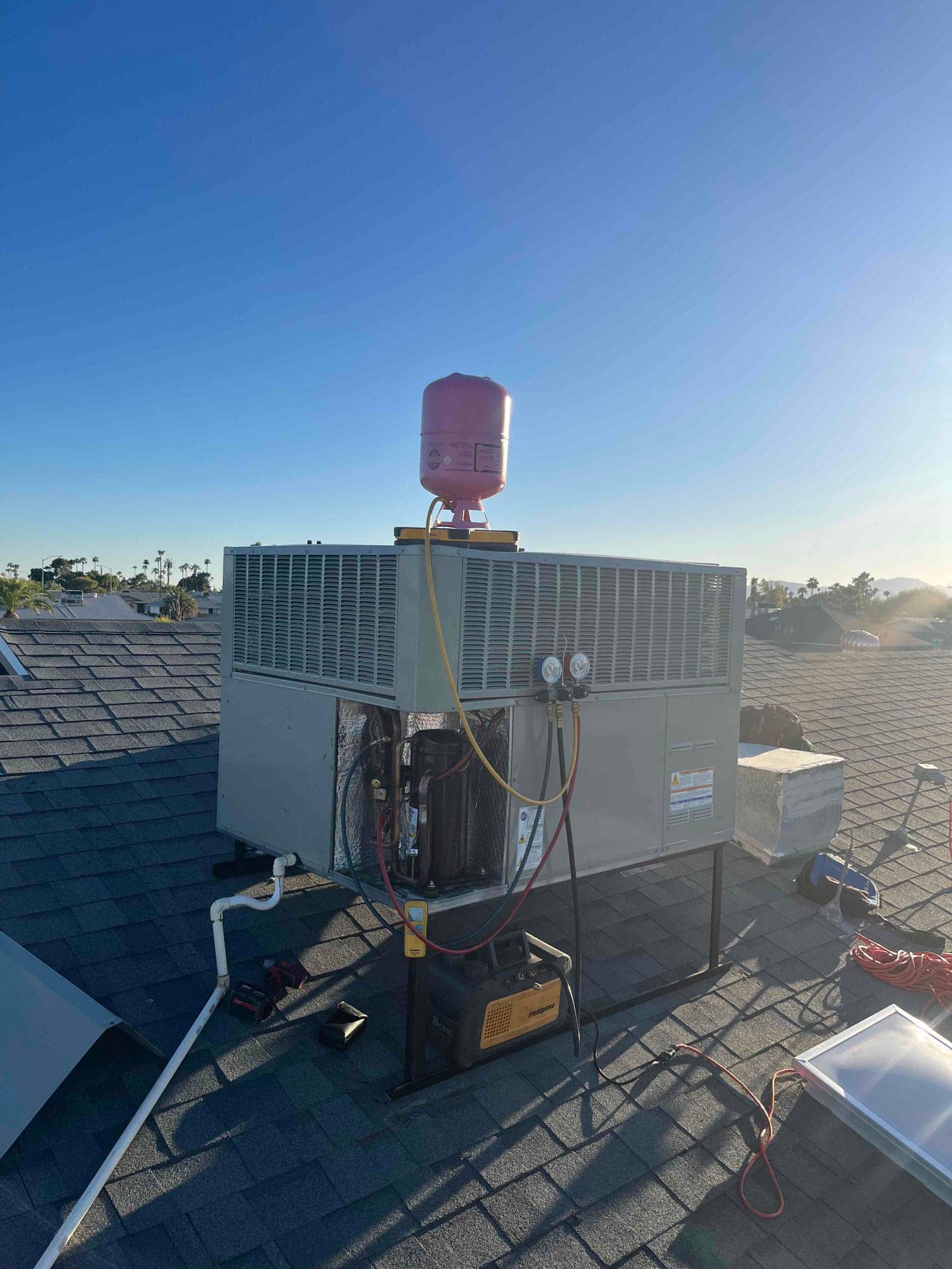 Rooftop package unit with service panel open, manifold gauges connected, refrigerant cylinder above, and vacuum pump on the roof during service.