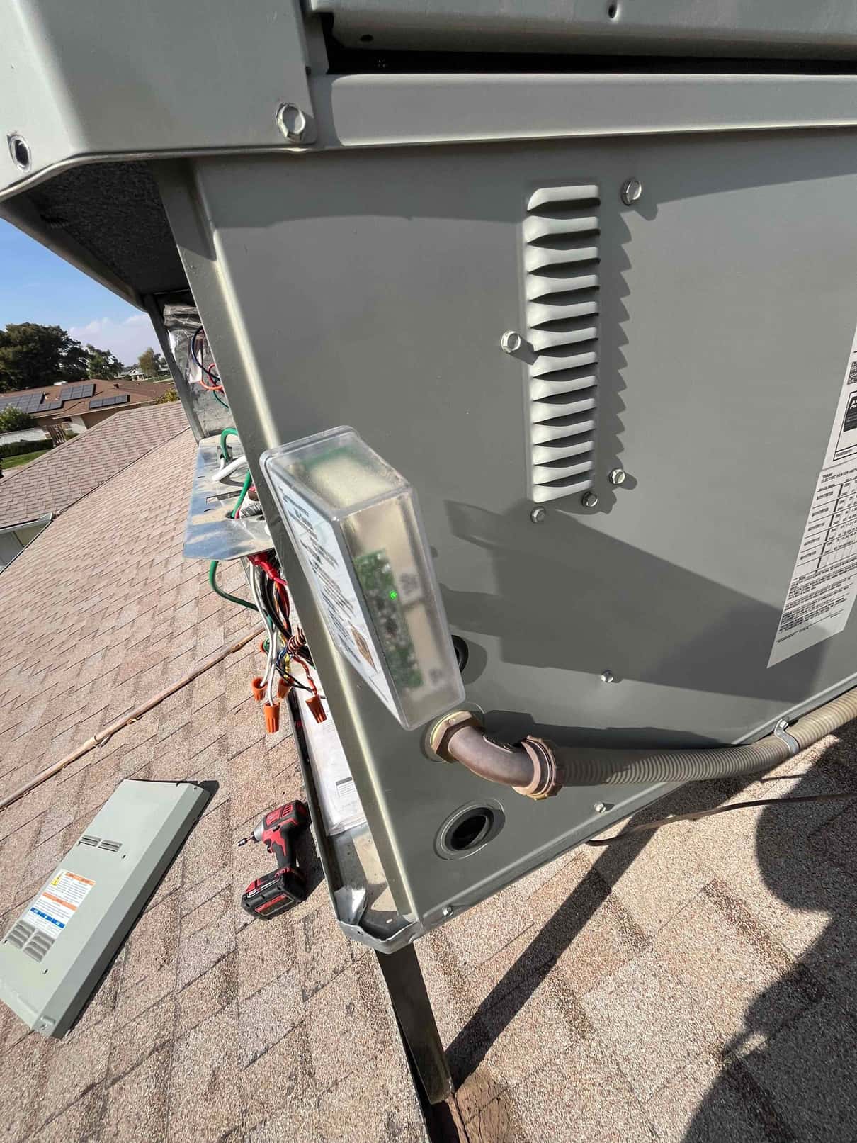 Rooftop packaged HVAC unit with access panel removed, showing wiring, condensate drain connection, flexible conduit, and an attached electronic module with green LEDs.