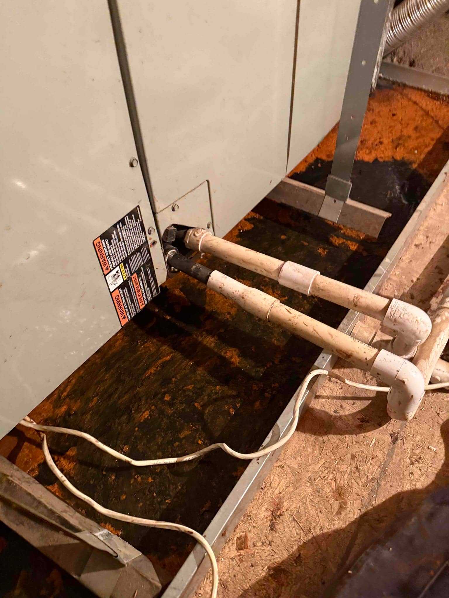 Air handler with PVC condensate drain lines above a metal drain pan containing standing water and rust-colored residue.