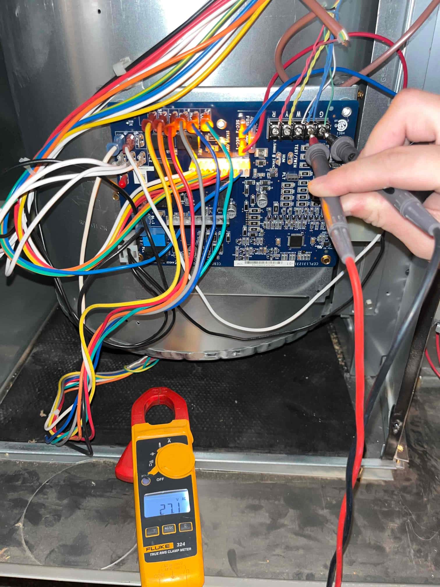 Control board with multiple colored wires; technician tests the terminal block with meter probes inside the HVAC unit cabinet.