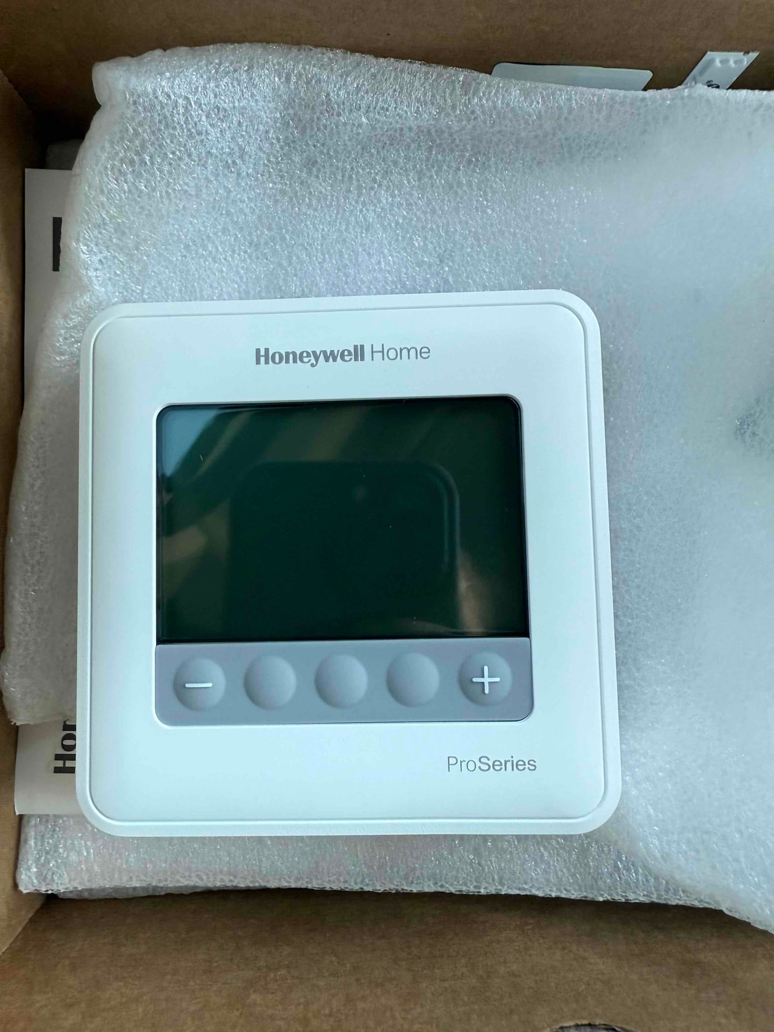 Digital wall thermostat in its packaging, showing a blank screen and five gray control buttons.