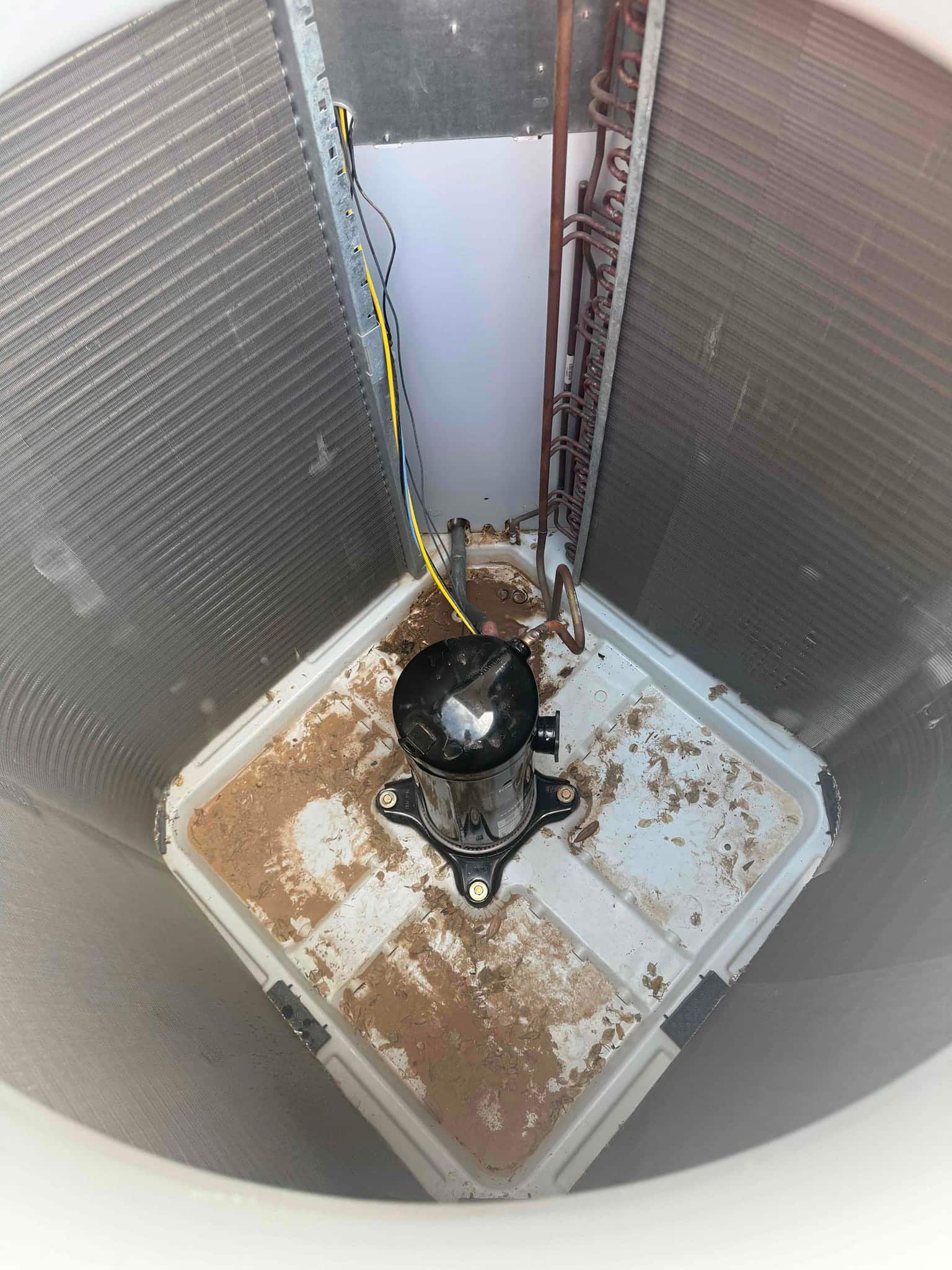Open outdoor condenser cabinet showing surrounding coil fins, base pan with debris, loose copper tubing, and wires.