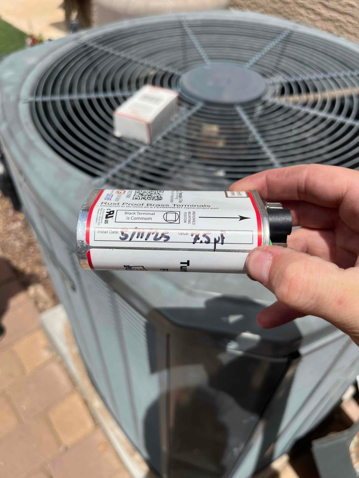 Technician holds a metal capacitor with terminal diagram and handwritten 45 μF marking in front of the outdoor condenser.